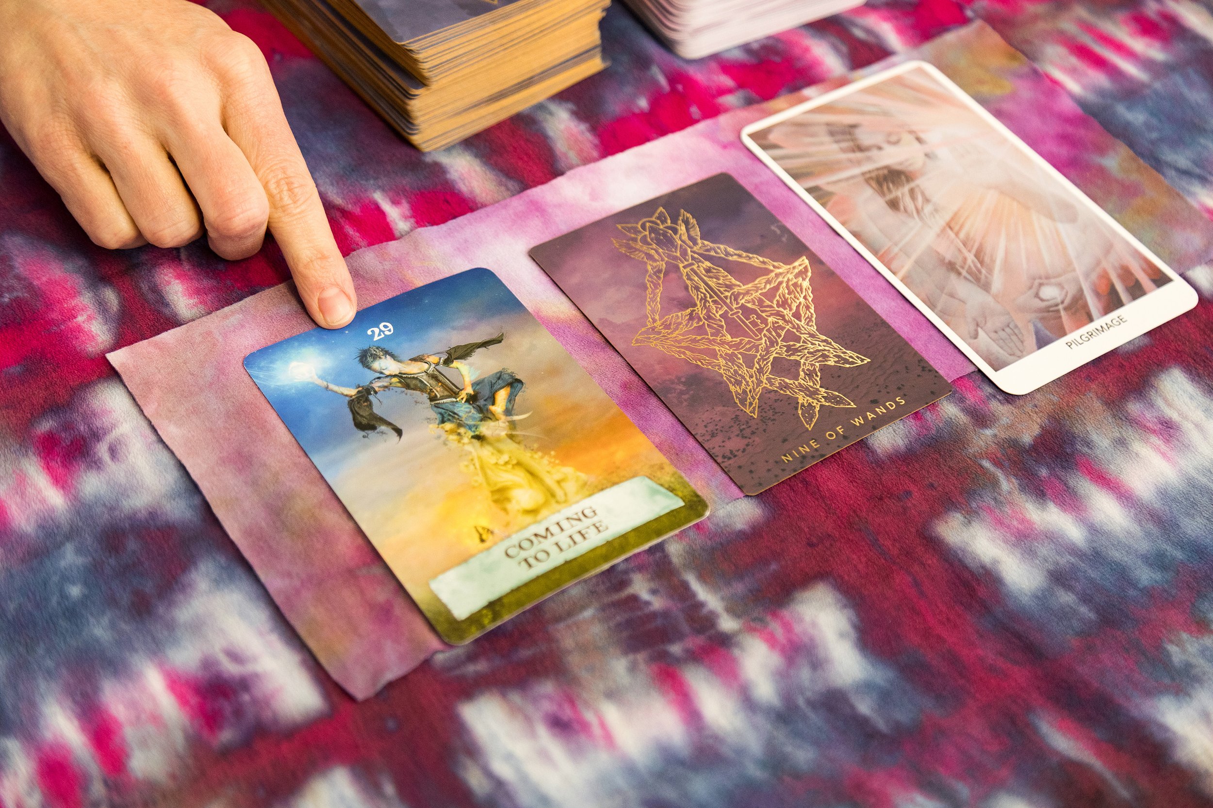 A person points to a tarot card titled 'Coming to Life' with an illustration of a female figure, next to other tarot or oracle cards placed on a colorful, tie-dye cloth.