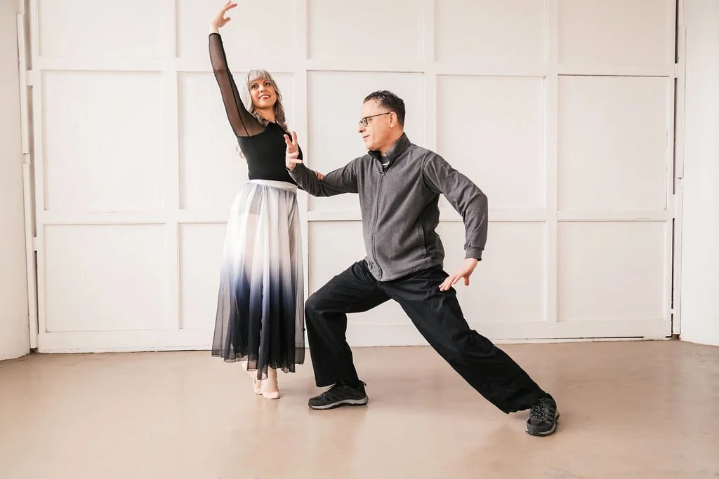 A woman in a ballroom dance pose wearing a black top and long flowing skirt, and a man in casual athletic clothes in a dance stance, practicing dance together indoors.