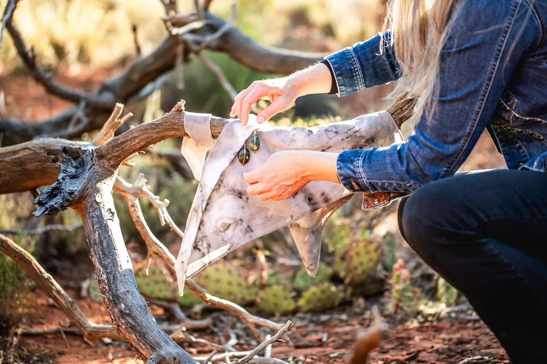 A person placing jewelry on a piece of fabric draped over a fallen tree branch in a natural outdoor setting.