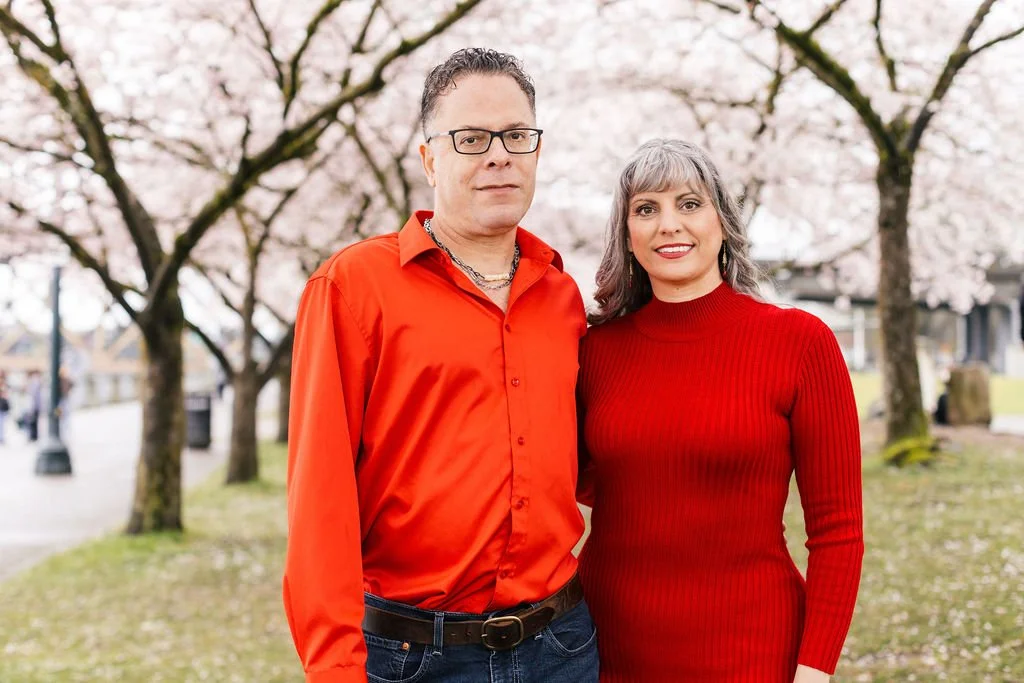 A man in a red shirt and a woman in a red dress standing outdoors with blooming cherry blossom trees in the background.