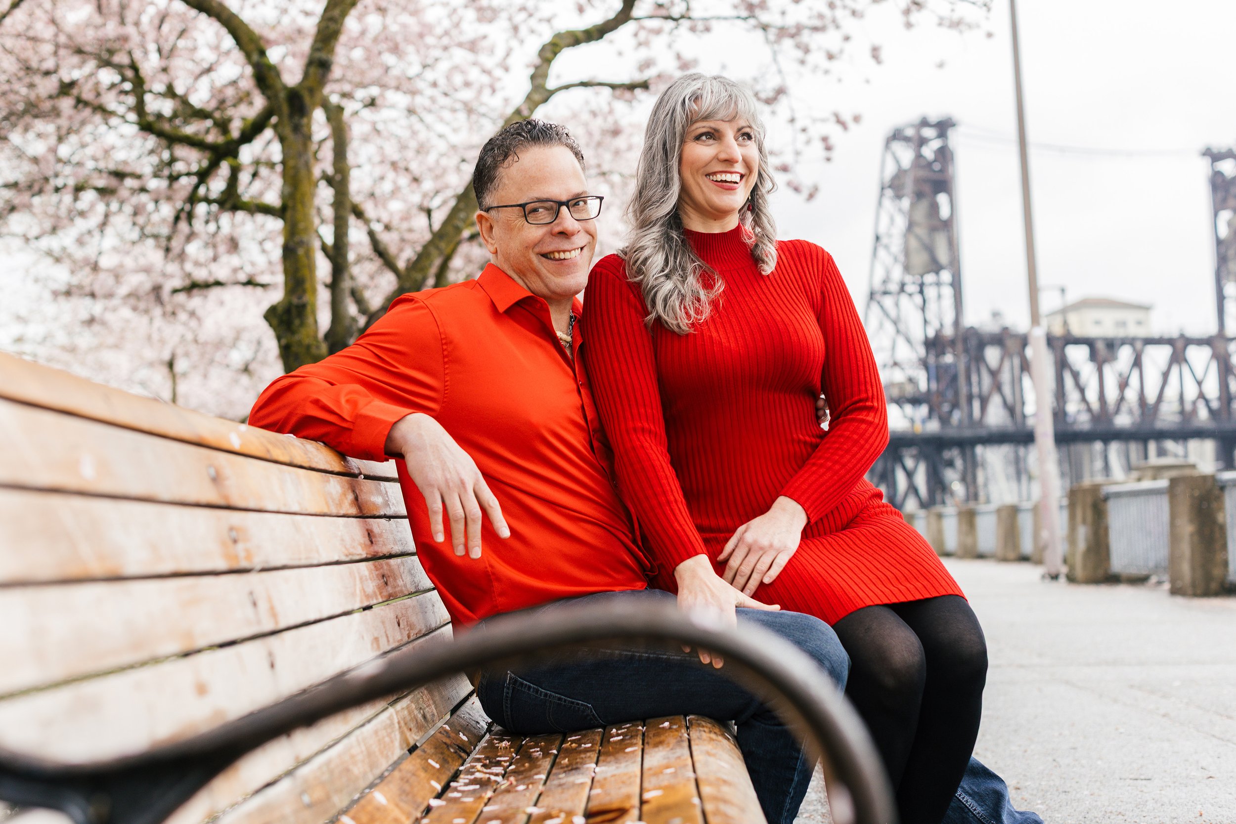 A smiling middle-aged man with glasses and a woman with gray hair seated on a wooden park bench with cherry blossoms and bridge in the background.