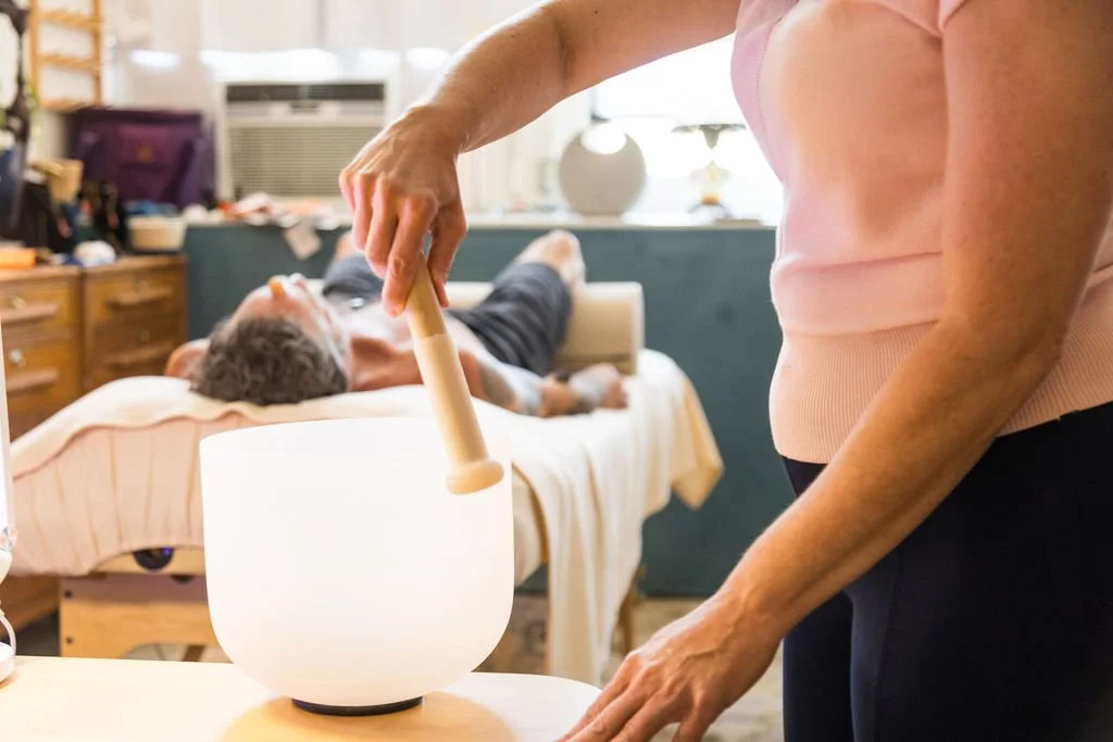 Person playing singing bowl while lying on a massage table, with another person in the background receiving a massage.