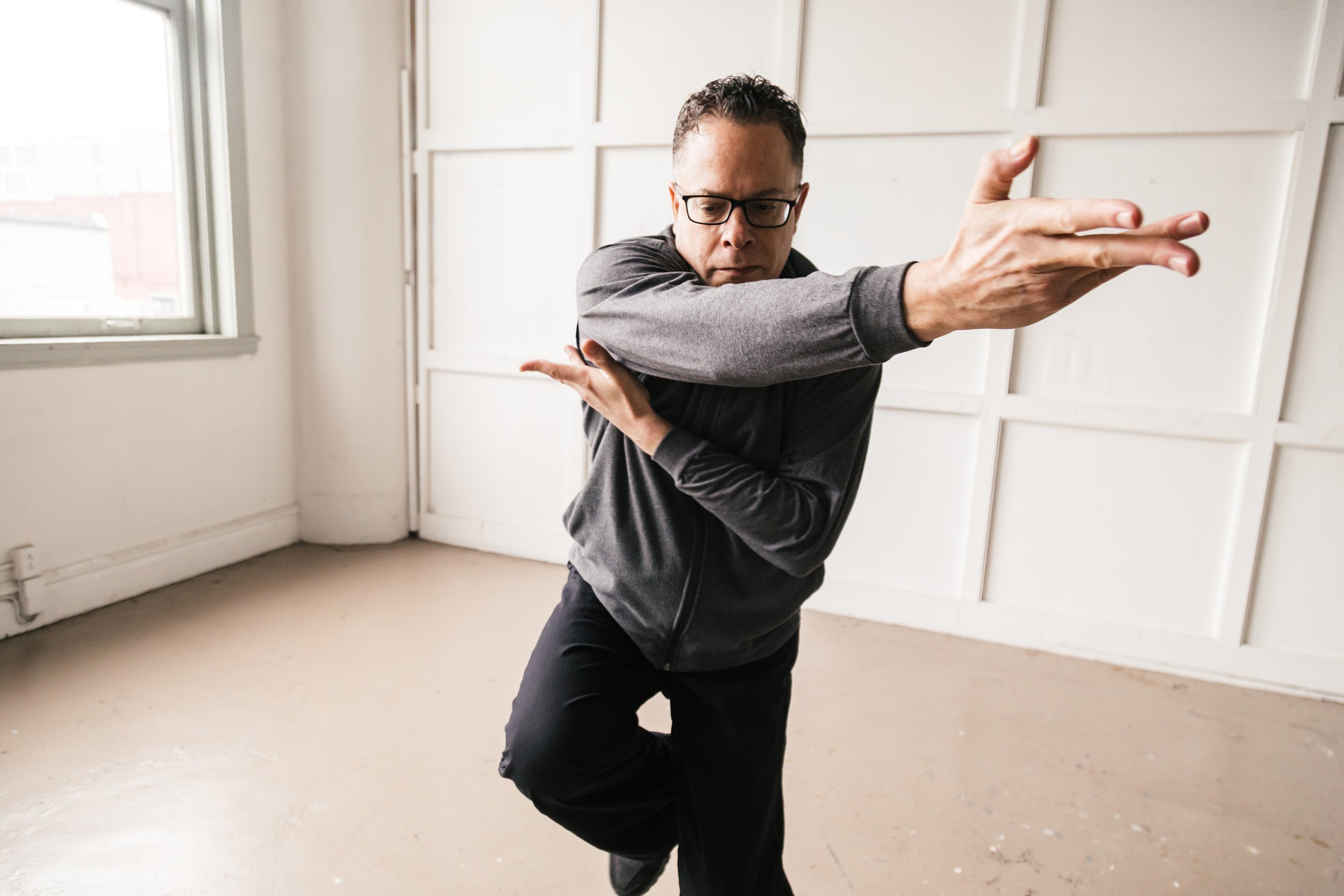A man with glasses and dark hair, wearing a gray zip-up jacket, is practicing a martial arts pose indoors. He is standing with his left arm extended straight forward, his right arm bent across his chest, and his right hand near his face. The room has