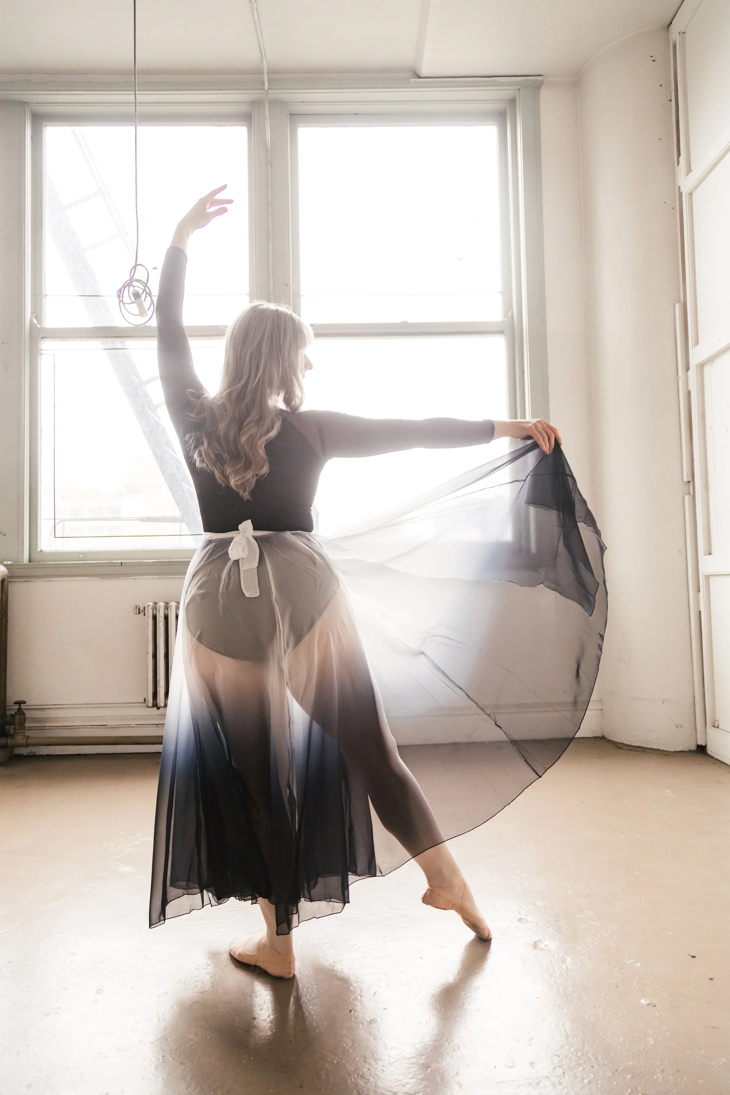 A ballerina in a black and white dress practicing ballet in a sunlit studio with large windows and a concrete floor.