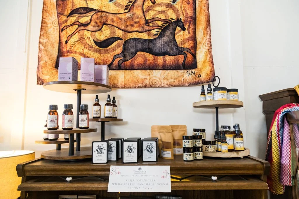 Display of various wellness products including bottles of oil, candles, and skincare products on wooden shelves in a cozy boutique, with a tapestry featuring horse artwork hanging on the wall behind.
