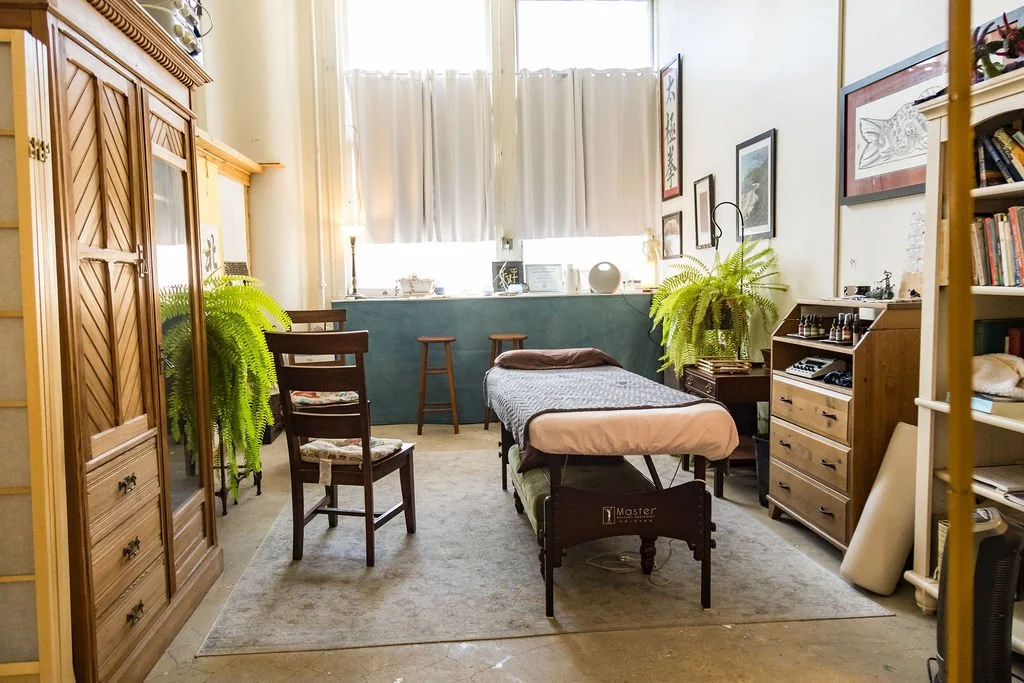 A massage room with a massage table, wooden chairs, large windows with white curtains, potted plants, and various decor on the walls and shelves.