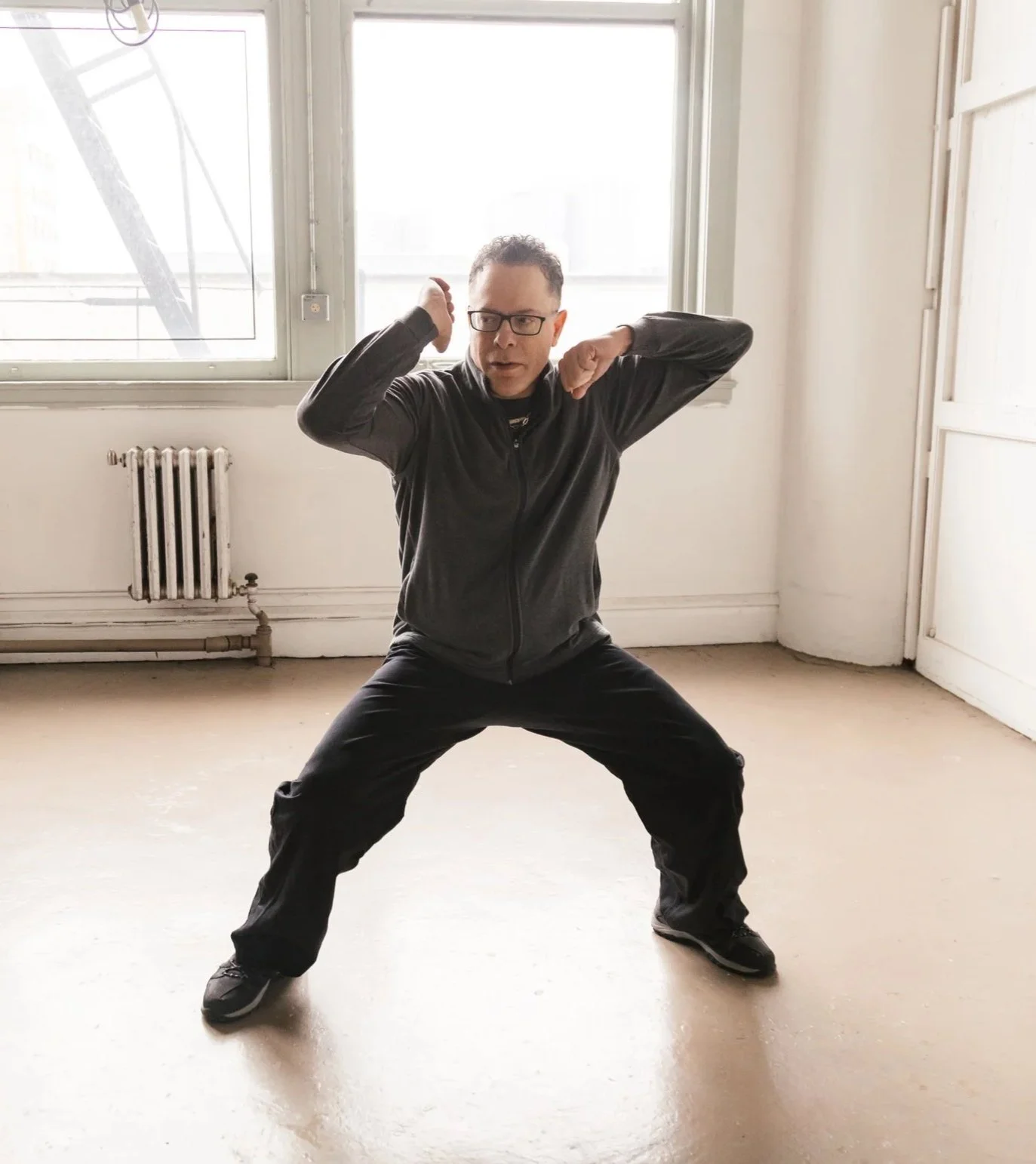 A man wearing glasses and a black jacket practicing a martial arts pose in a room with large windows and a radiator.
