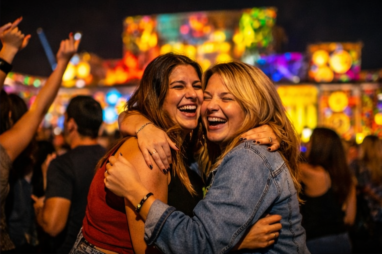 Two women hugging and laughing at an outdoor concert or festival at night, with a colorful, illuminated stage in the background and a crowd around them.