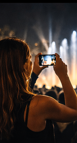 A woman taking a photo with her smartphone at a concert or event, with stage lights in the background.