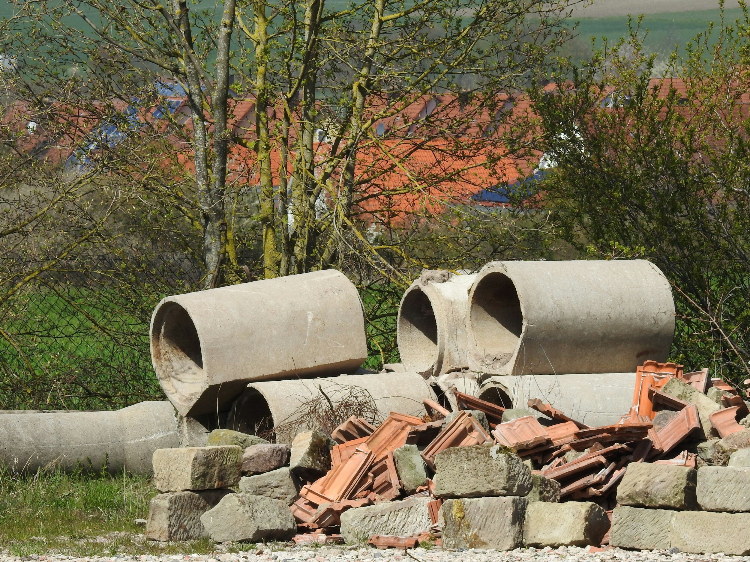 Construction waste including large concrete pipes, bricks, and stones stacked outdoors with trees and rooftops in the background.
