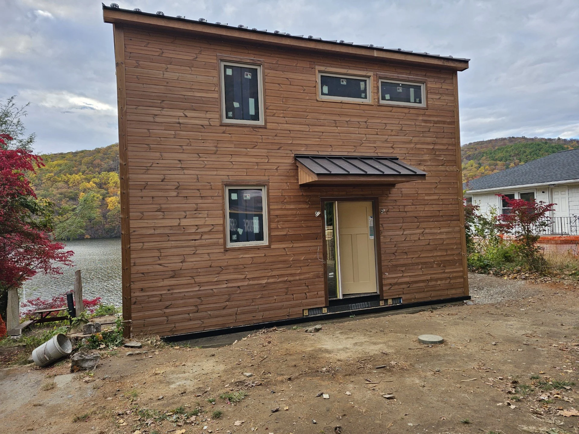 Under construction wooden house with a few small windows, a partially open door, and a small overhang above the door, situated near a river with trees and hills in the background.