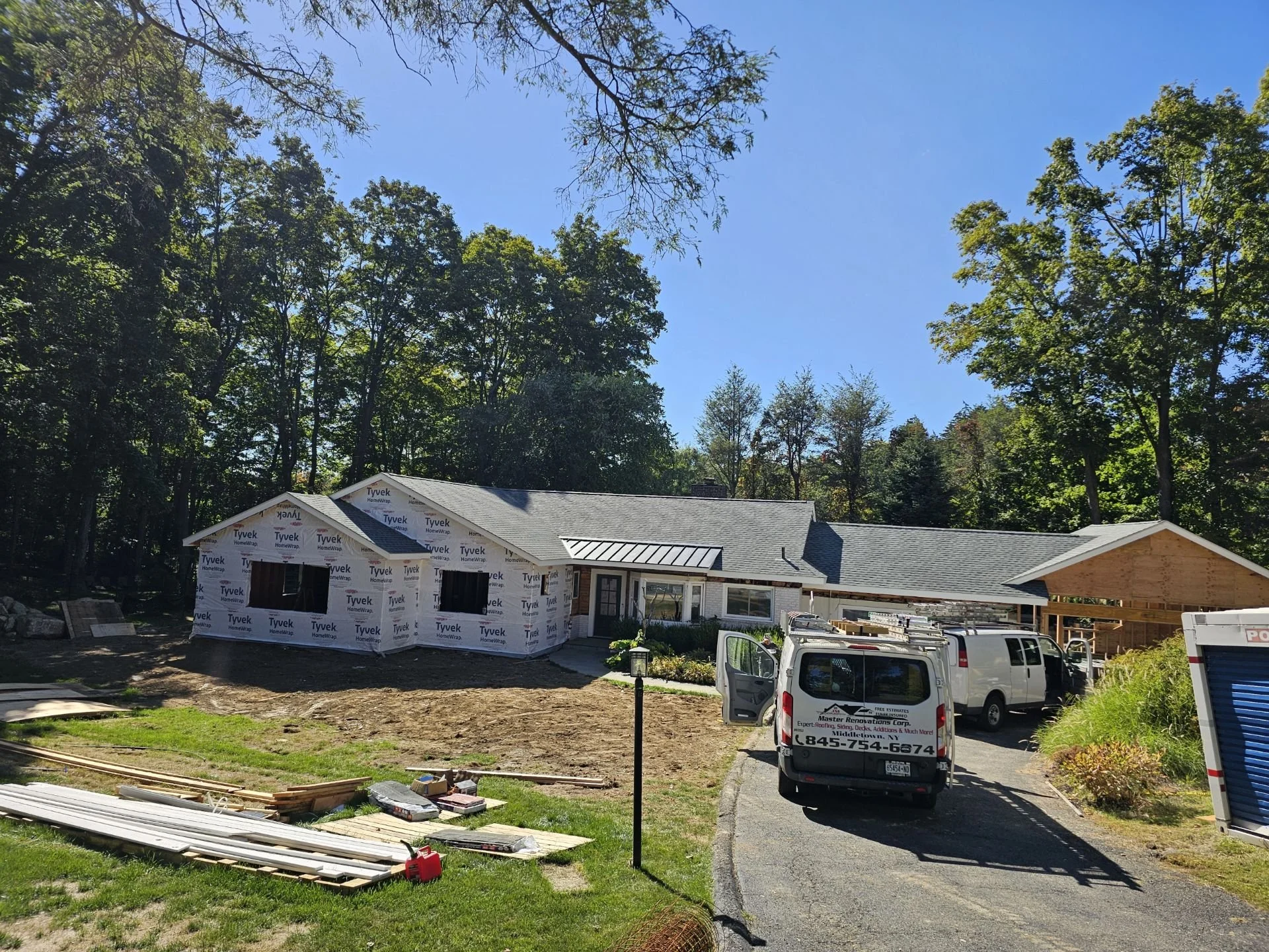 A house under construction with roofing work in progress, parked vans on the driveway, and construction materials on the lawn during a sunny day with trees in the background.