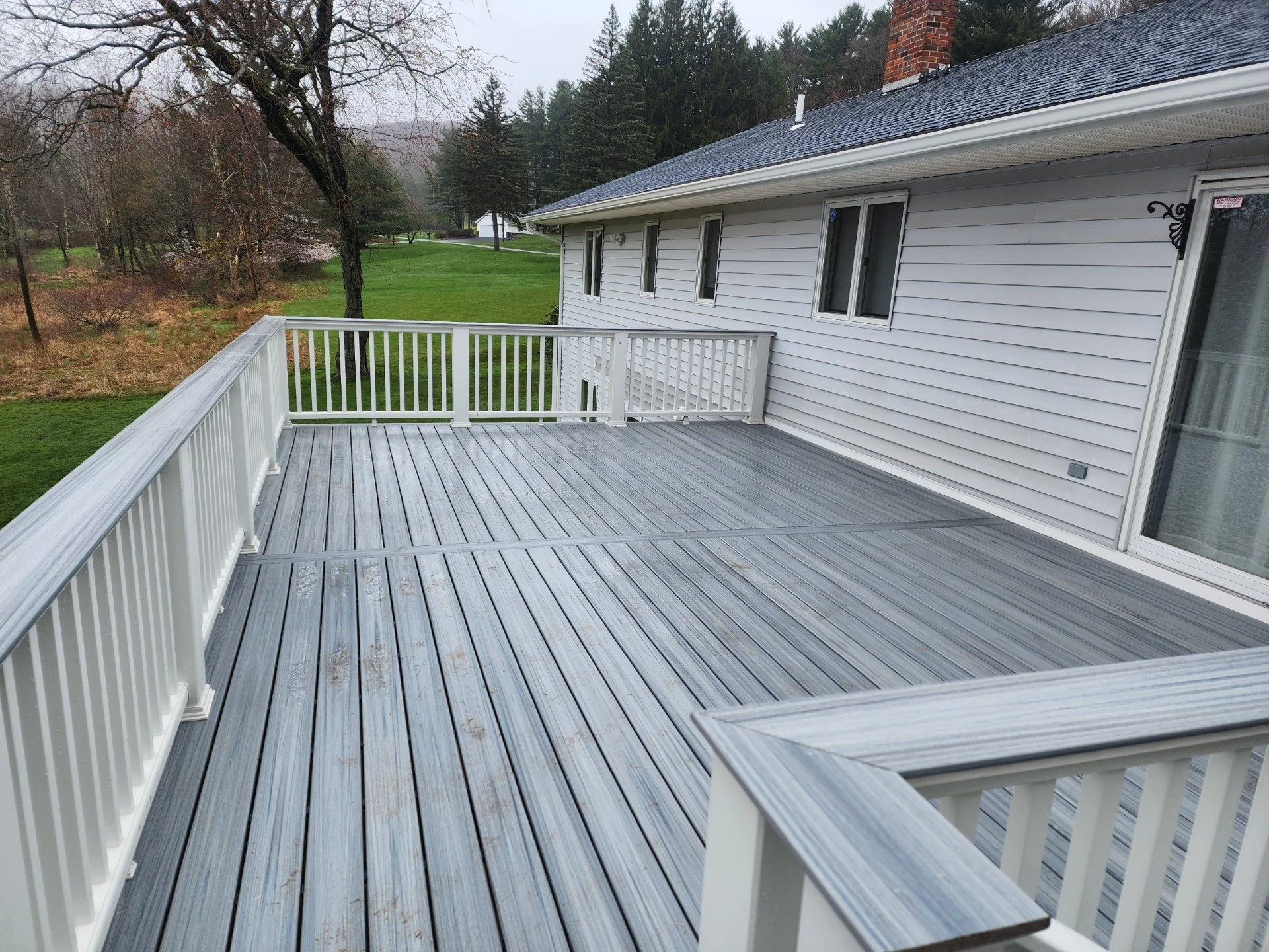 Empty wooden deck attached to a white house with white railing, overlooking a green lawn and trees in the distance on an overcast day.