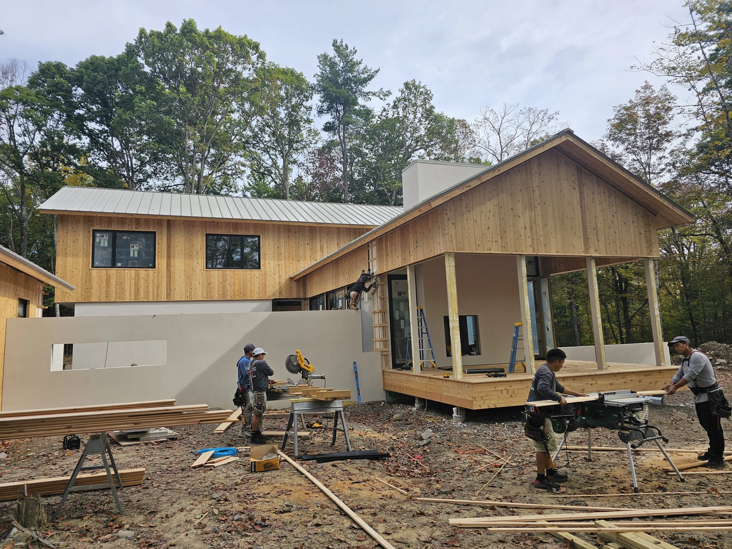Construction workers building a two-story house with wooden exterior walls, some work on the porch and others operate saws and tools. The house is located in a wooded area under a cloudy sky.