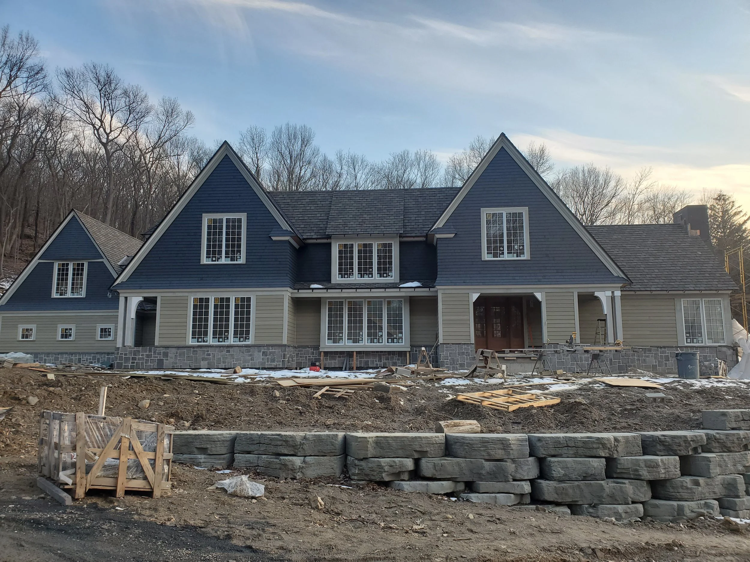 Under construction large house with blue and beige siding and multiple gabled roofs, surrounded by construction materials and debris, with leafless trees in the background.