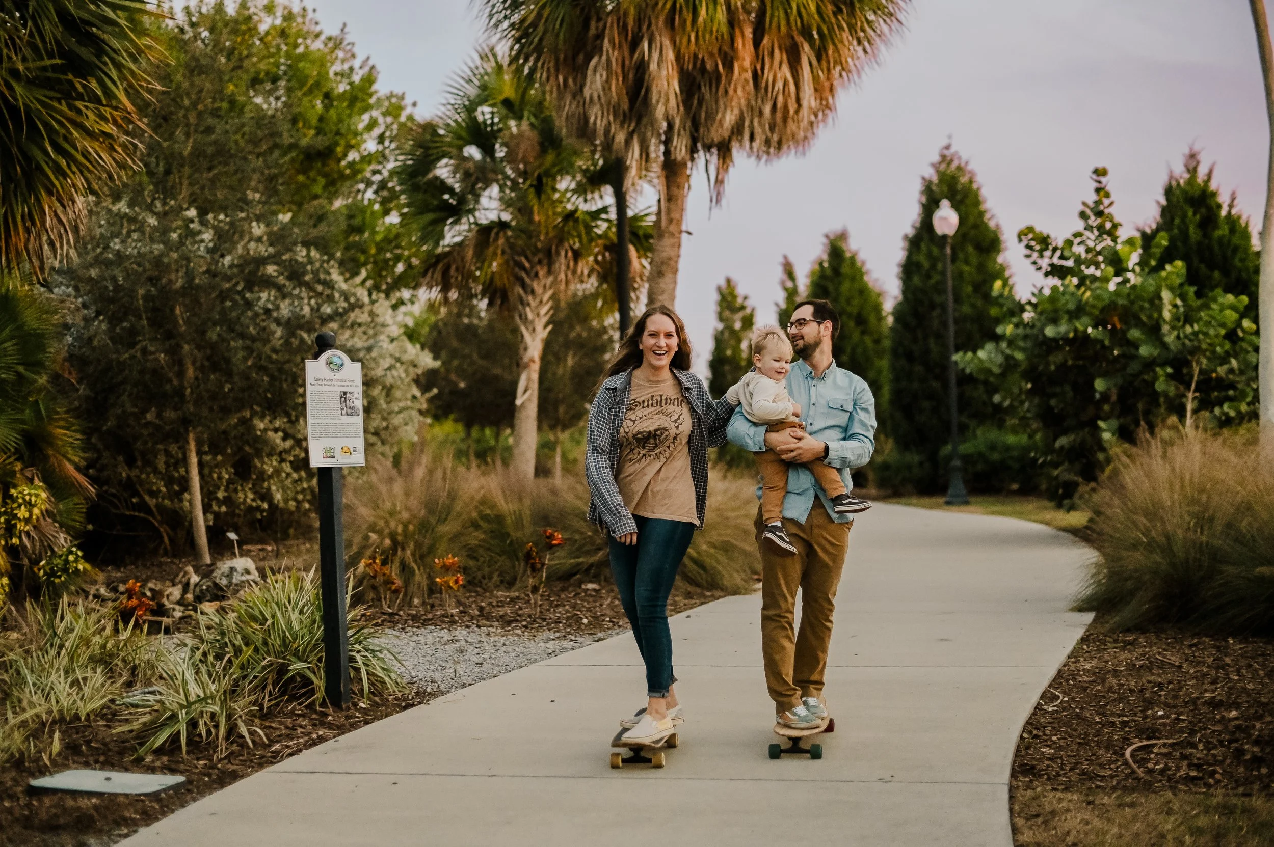 A family of three enjoying a walk and skateboarding along a paved path in a park with lush greenery and palm trees during the evening.