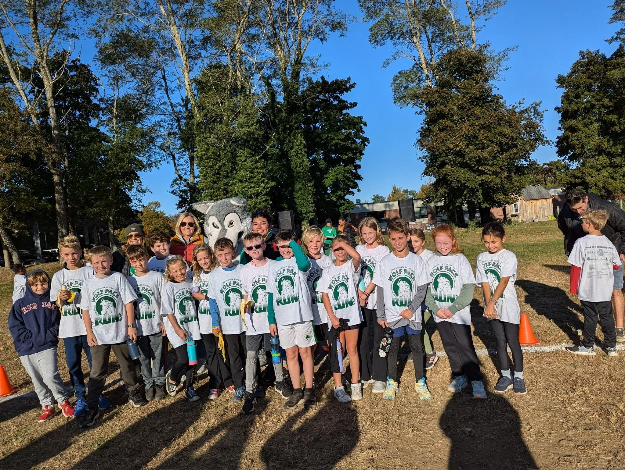A large group of children and adults gathered outdoors for a group photo at a school event, with trees and a building in the background. Some children are wearing white T-shirts with the logo 'Wolf Pack' and a wolf mascot.