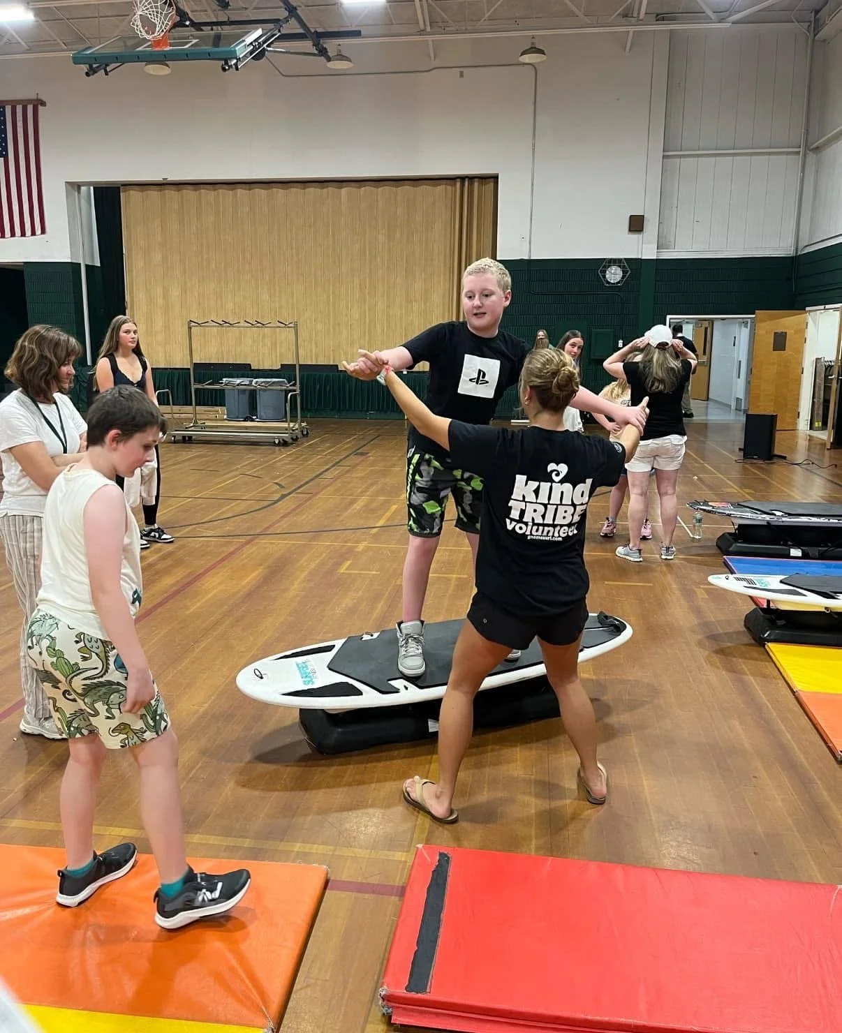 A group of children and a volunteer practicing surfing indoors on a makeshift surfboard with a padded base in a gymnasium. The scene involves children standing on mats while the volunteer helps a boy balance on the surfboard.