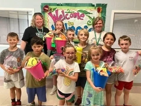 Group of children and two adults standing in front of a welcome sign, holding gift buckets and toys in an indoor setting.