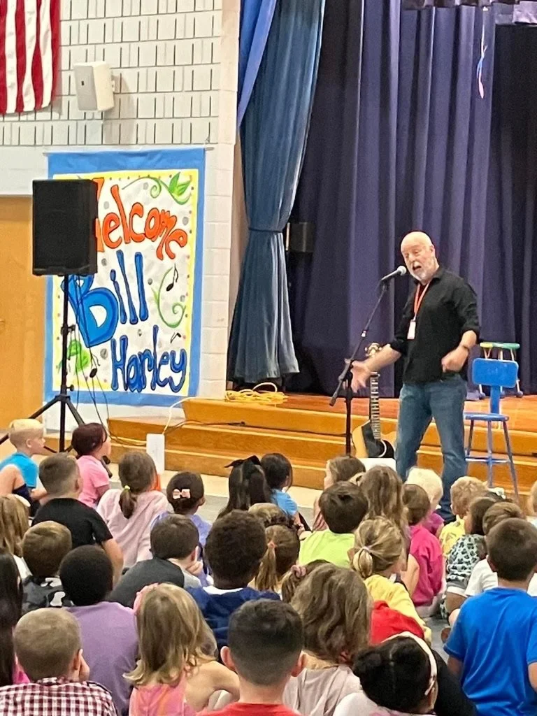 A man performing on stage with a guitar and microphone in front of a crowd of children at school assembly. A colorful welcome banner with the text "Welcome Bill Harley" is on the wall.