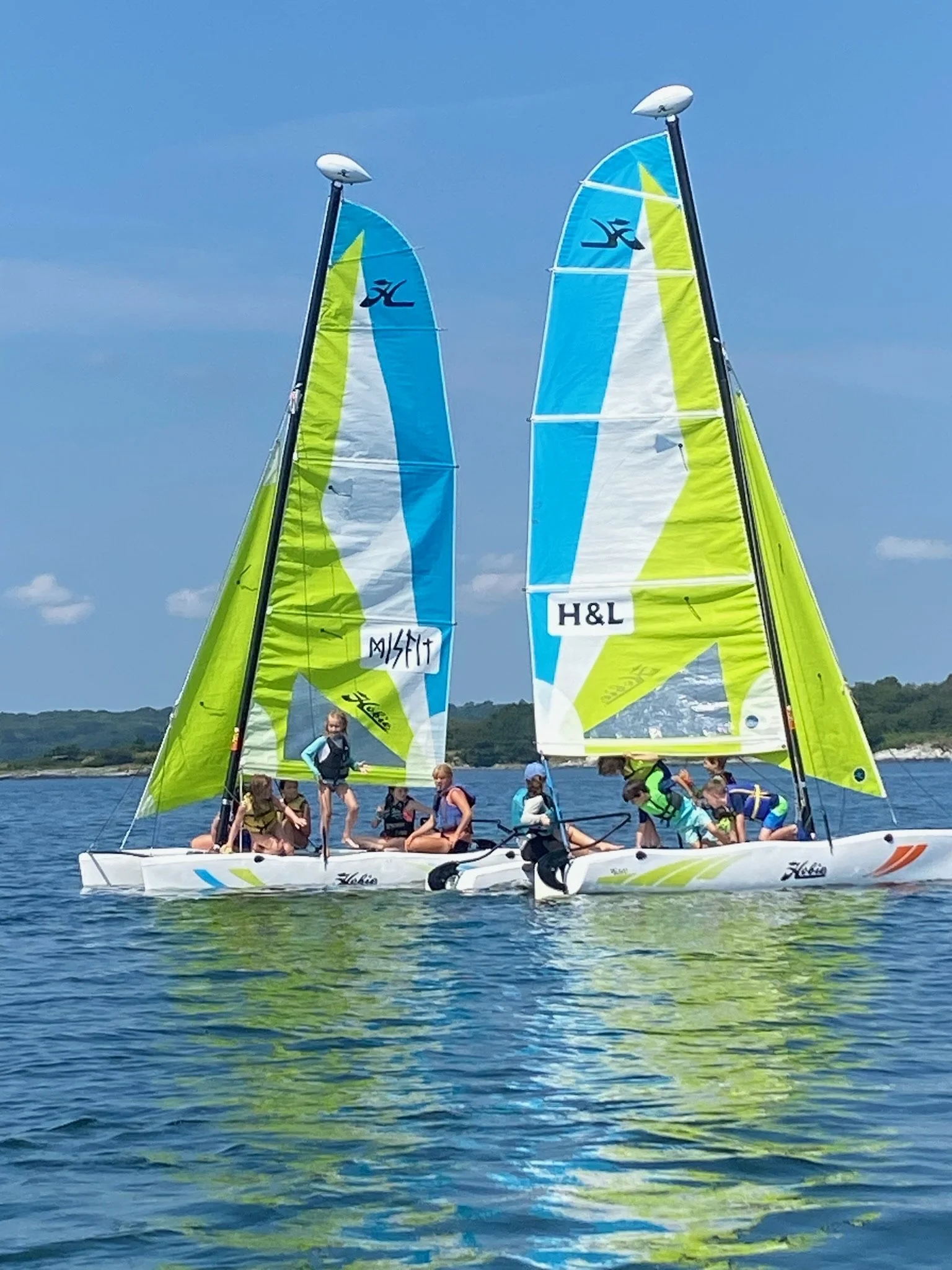 Children sailing on a bright, multi-colored sailboat with yellow, blue, and white sails on a lake under a partly cloudy sky.