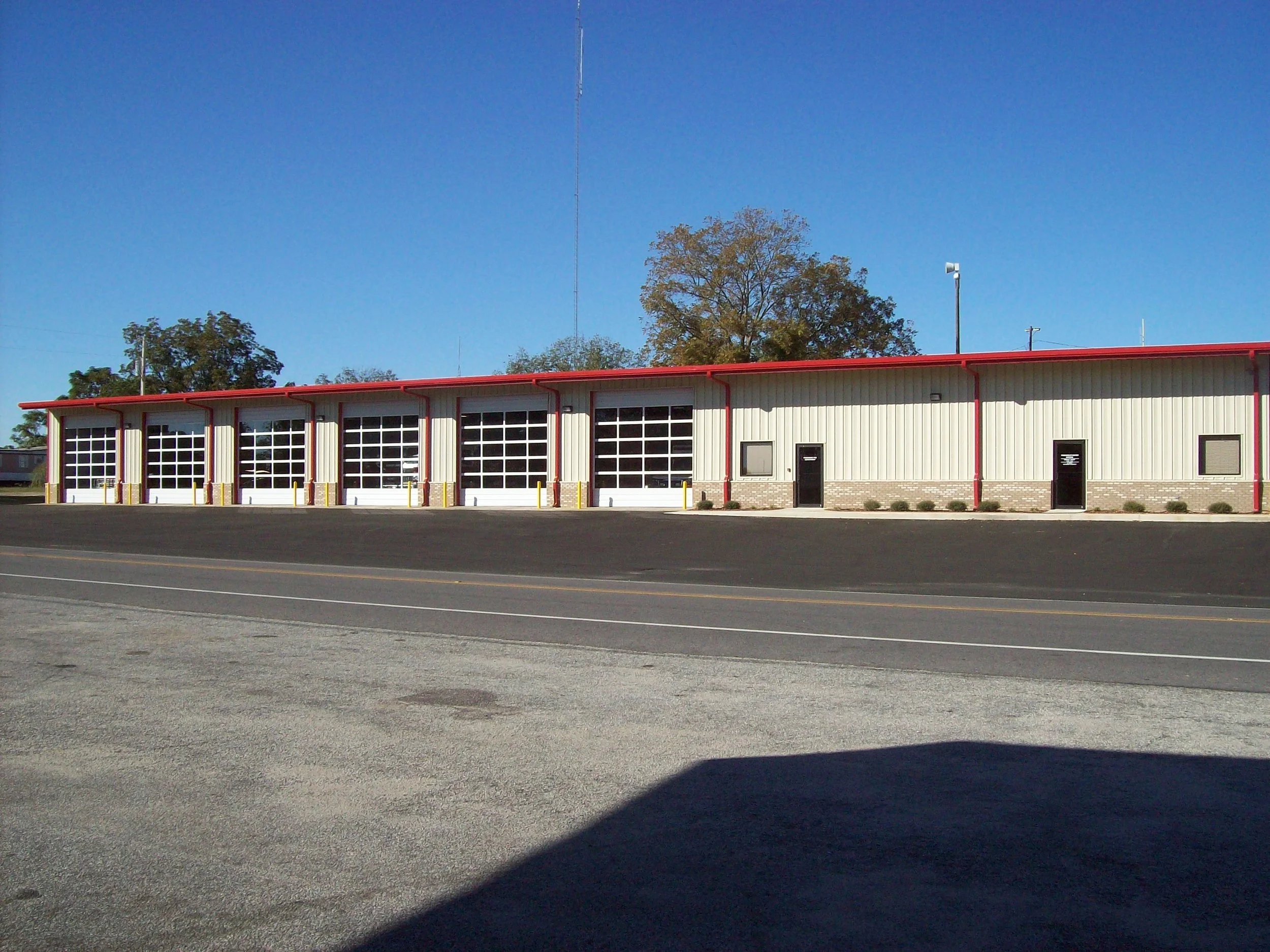 A large building with multiple garage doors, two black doors, beige exterior walls, red trims, and a paved parking area in front, under a blue sky with trees in the background.