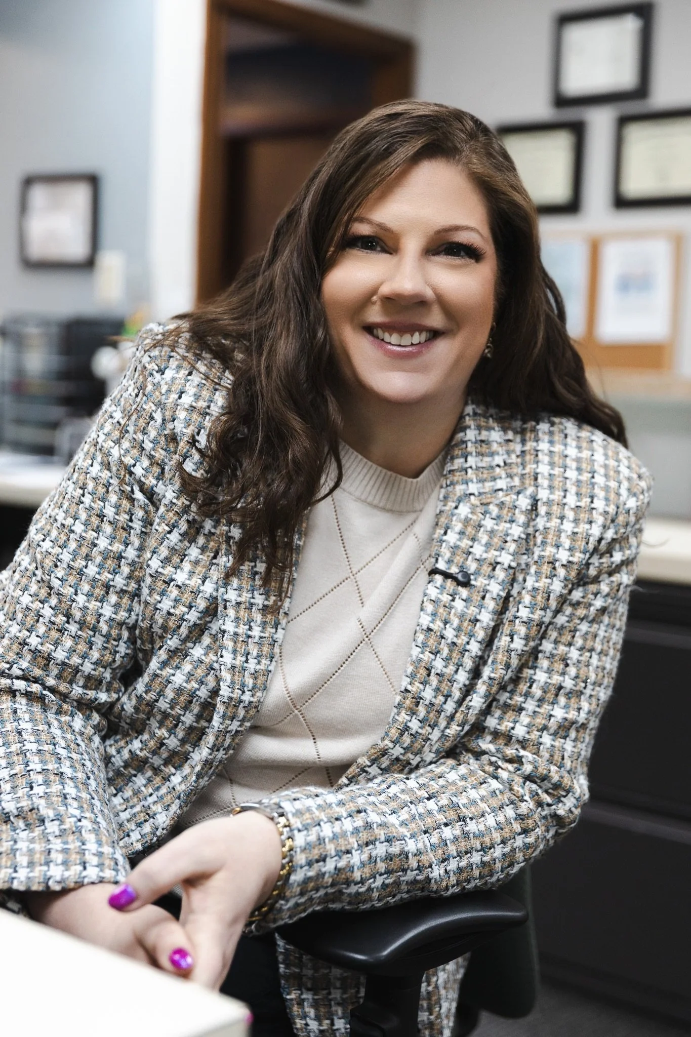 A woman with long brown hair and a checkered blazer, smiling in an office setting.