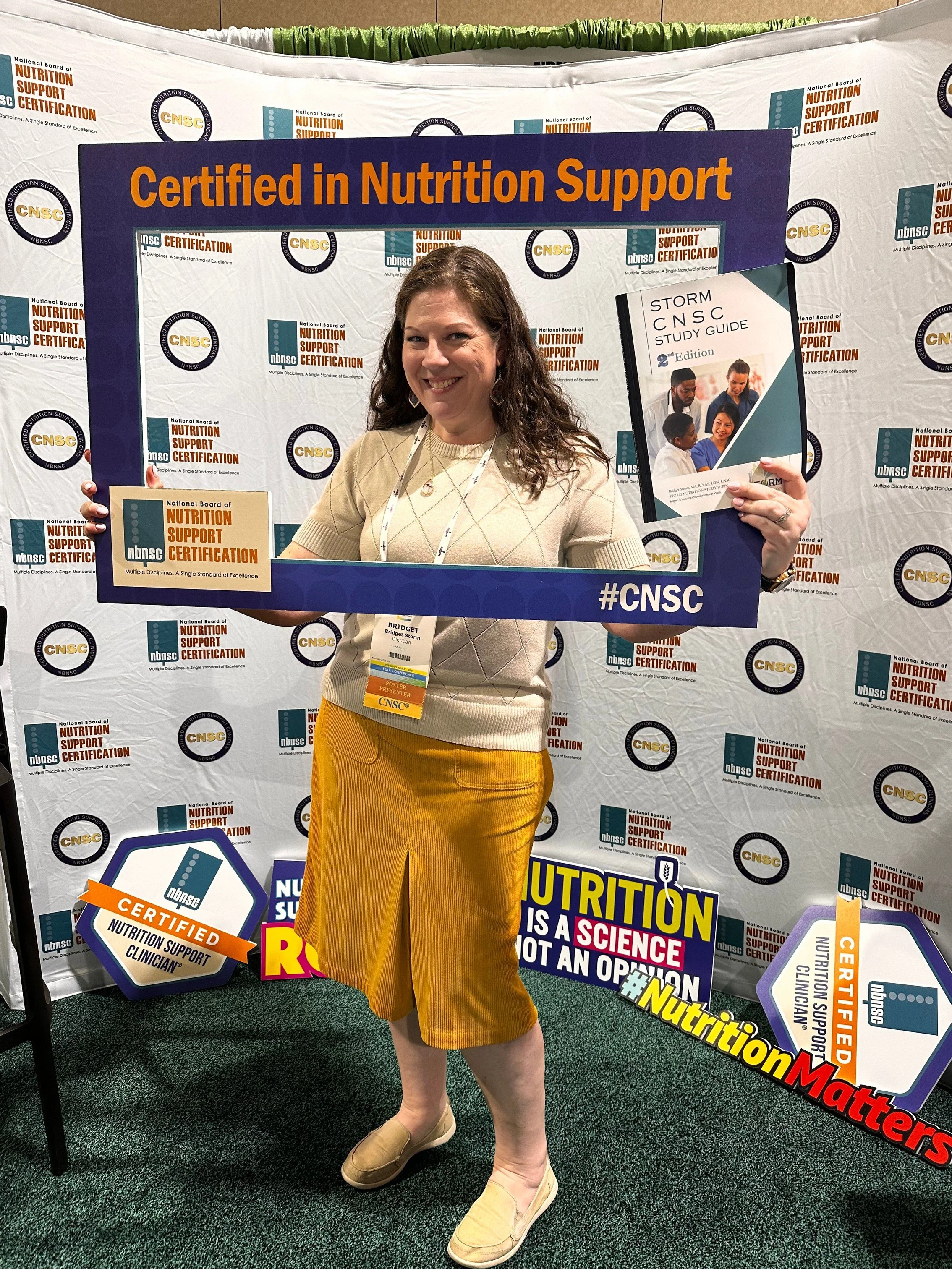 Woman standing inside a photo booth at a nutrition support certification event, holding a study guide and smiling. Background features banners and signs about certified nutrition support clinicians. She is wearing a beige sweater, yellow skirt, and beige shoes.