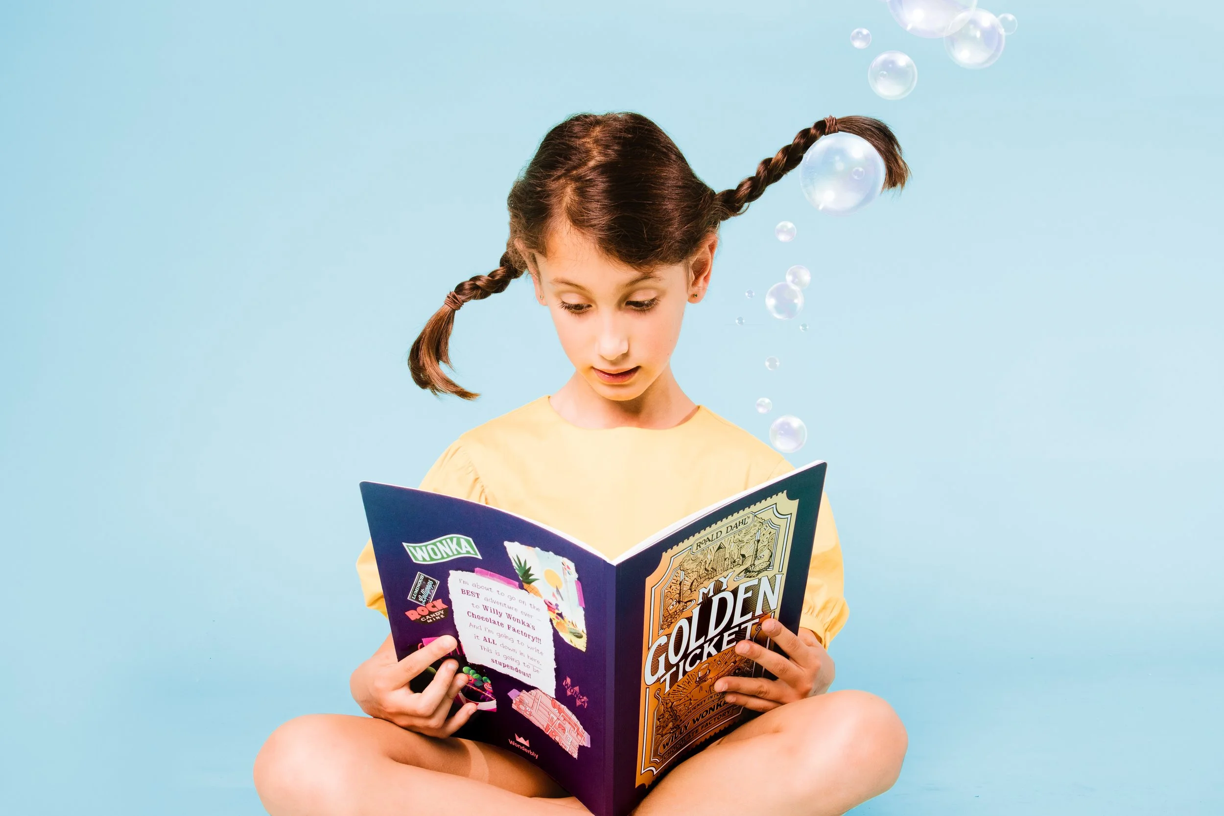 A young girl with braided hair, wearing a yellow shirt, sitting cross-legged against a blue background, reading a colorful book titled "Golden Ticket."