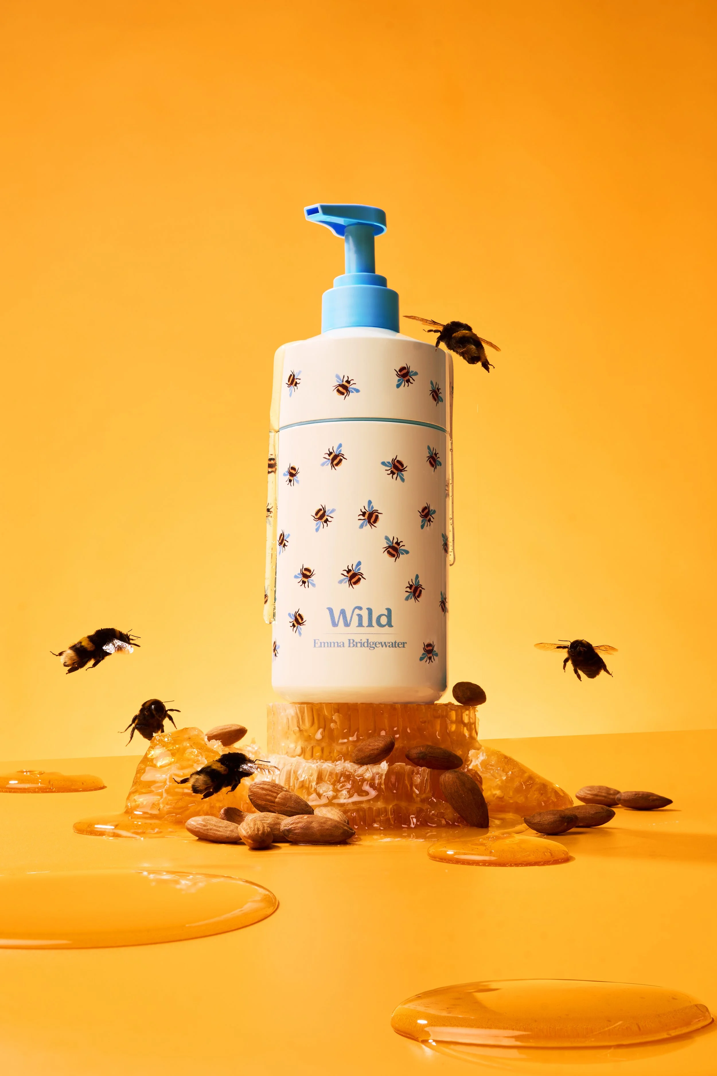 A white bottle of hand sanitizer with a blue pump, decorated with illustrations of bees and the word 'Wild' on it, placed on a piece of honeycomb. Several bees are flying around, and honey and pollen are scattered on a yellow background.