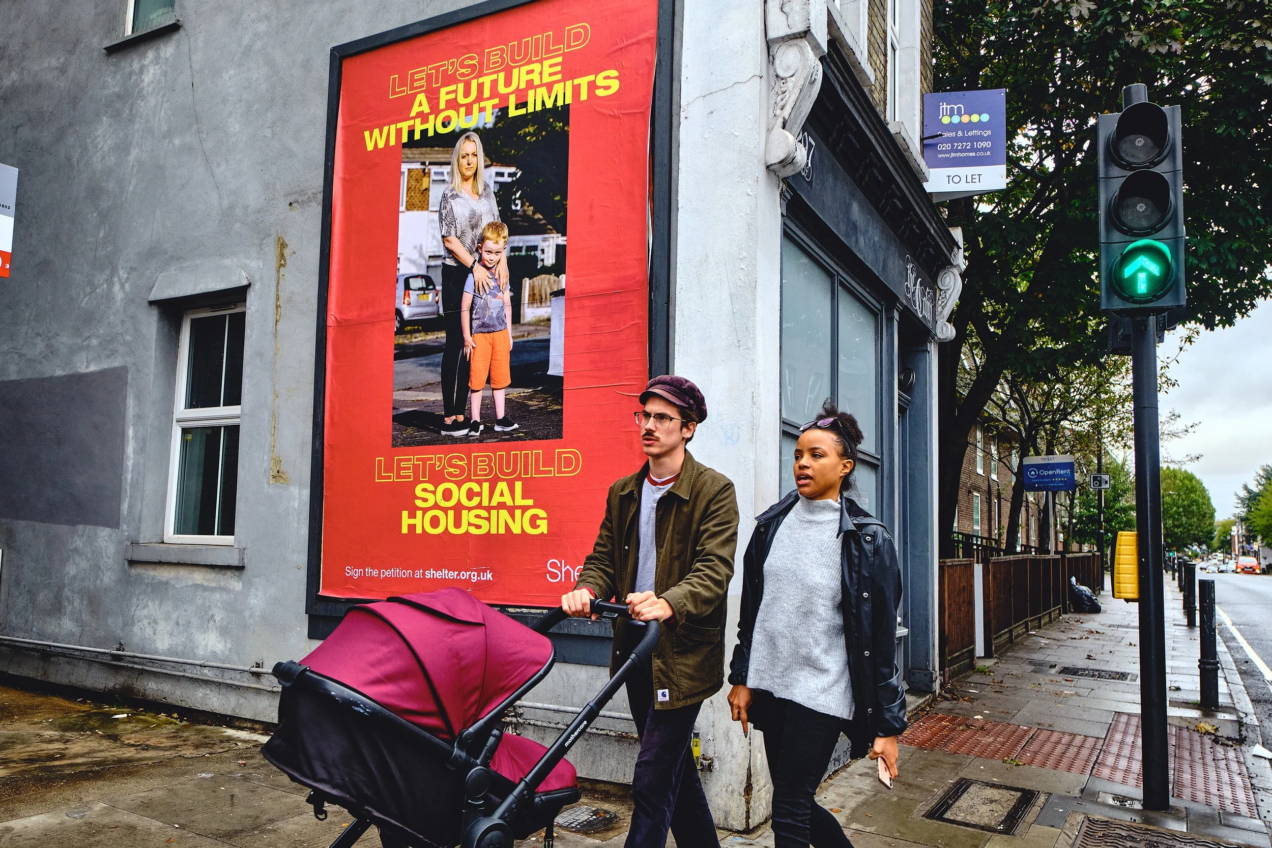 A man and woman walking on a sidewalk with a baby stroller. The man is wearing glasses and a green jacket, and the woman has her hair in a bun and is wearing a black leather jacket. They are passing a building with a large red poster that reads 'Let's Build a Future Without Limits' and 'Let's Build Social Housing'. There is a green traffic light showing an upward arrow, and trees are visible in the background.