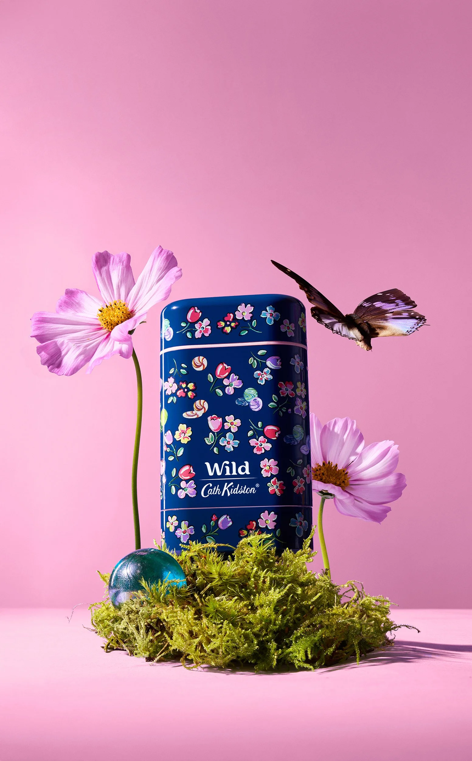 A floral patterned tin of Cath Kidston Wild perfume surrounded by pink cosmos flowers, green moss, a blue orb, and a butterfly with brown and white wings against a pink background.