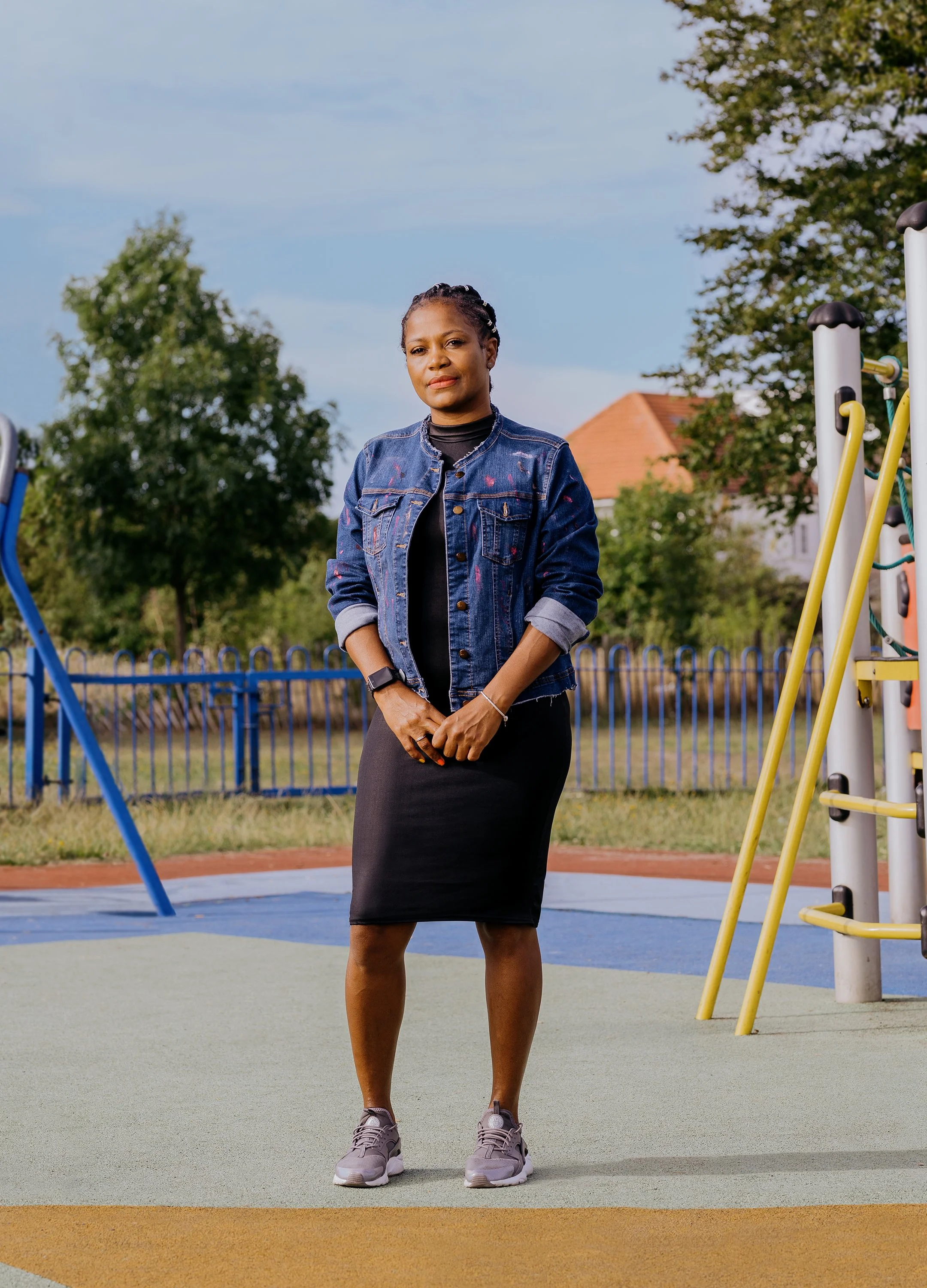 A woman standing on a playground surface, wearing a denim jacket, black dress, and gray sneakers, with trees and a house in the background.