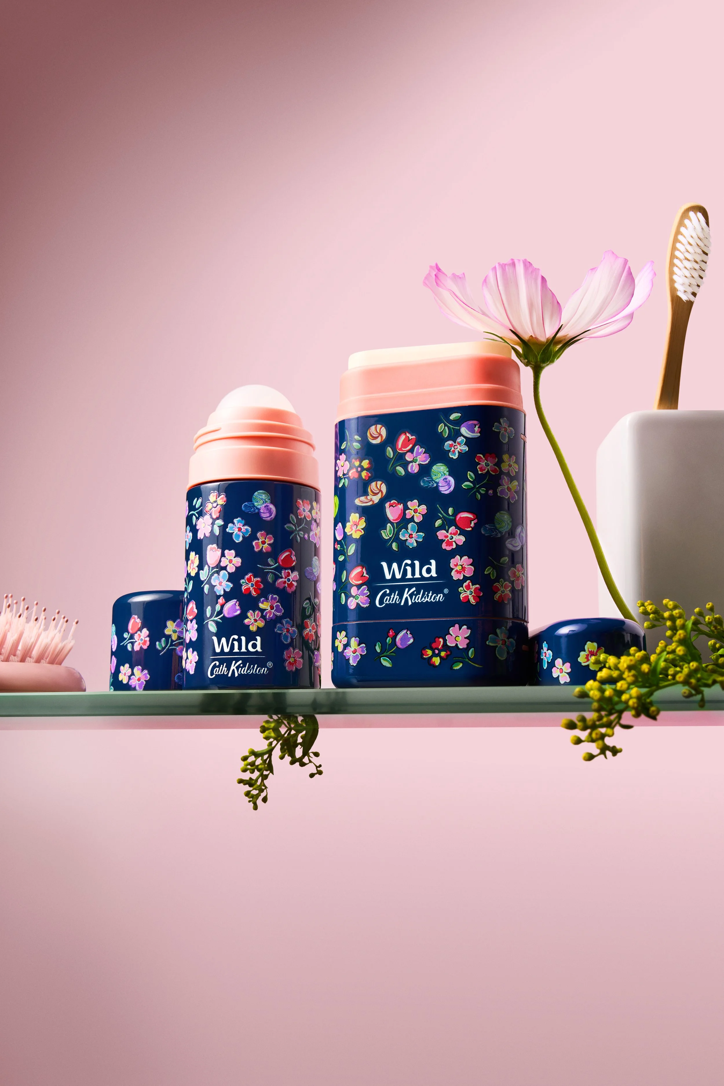 Two blue floral-patterned thermos bottles with pink caps and a pink flower on a glass shelf, along with a pink brush, a white container holding a toothbrush, and some yellow and green decorative plant elements against a pink background.