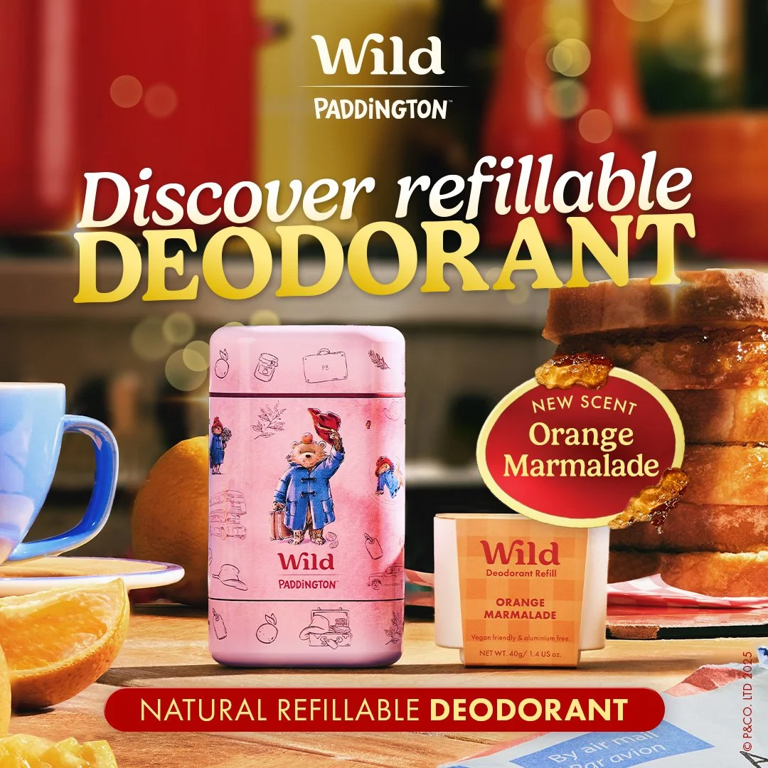 Packaging of Wild Paddington natural refillable deodorant with orange marmalade scent, featuring pink tin with Paddington bear illustration, display of orange slices, and a jar of marmalade on a wooden surface