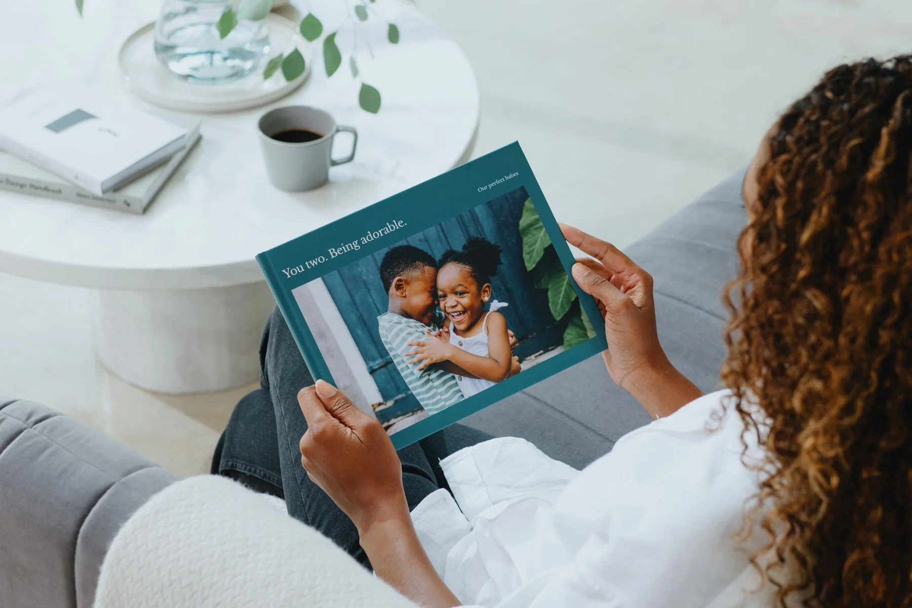 A woman sitting on a gray sofa holding a photo book with a picture of two children hugging, smiling, with the text 'You two. Being adorable.' on the cover.