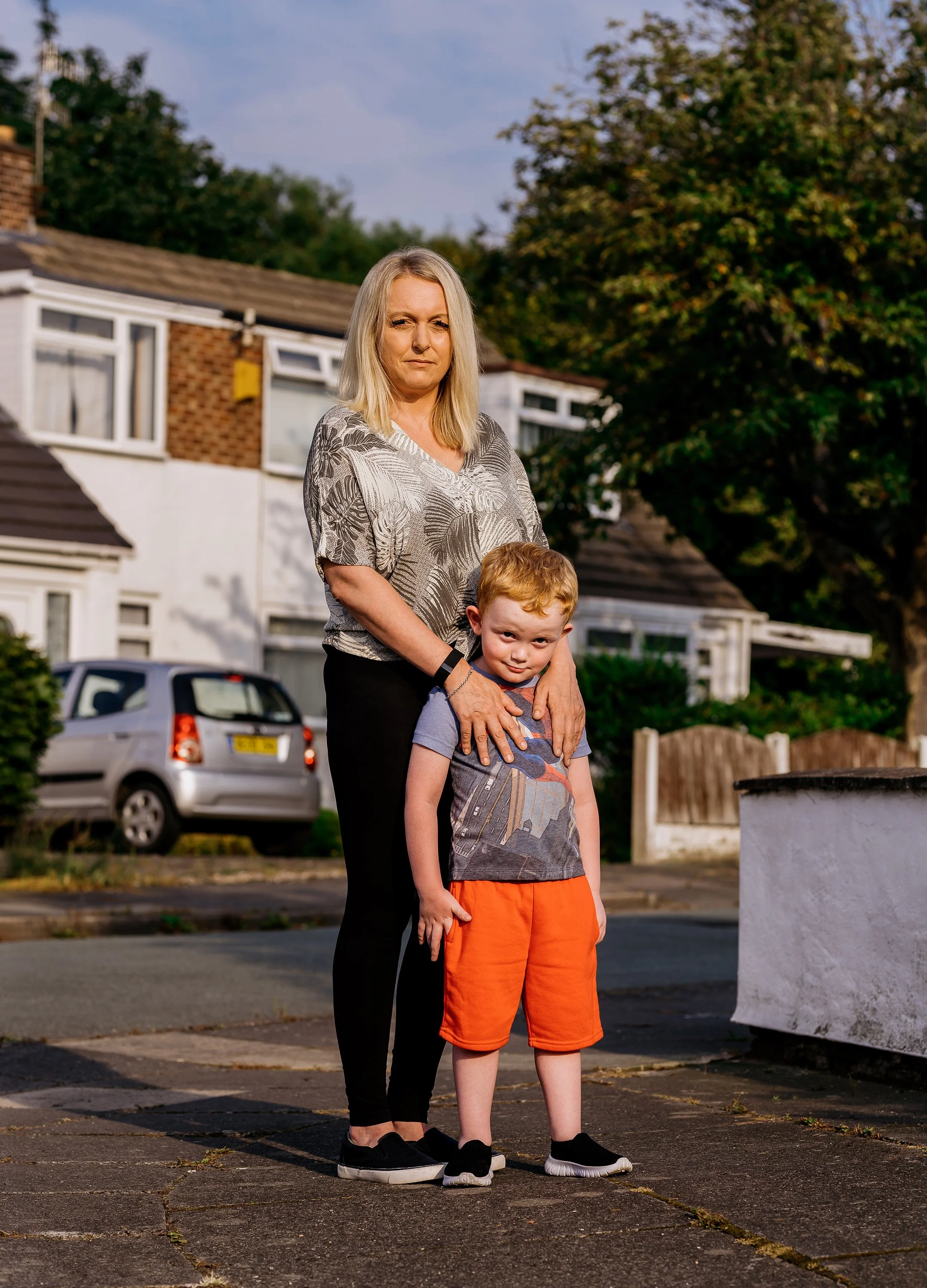 A woman and a young boy stand outdoors on a city street, with houses and parked cars in the background during daylight.