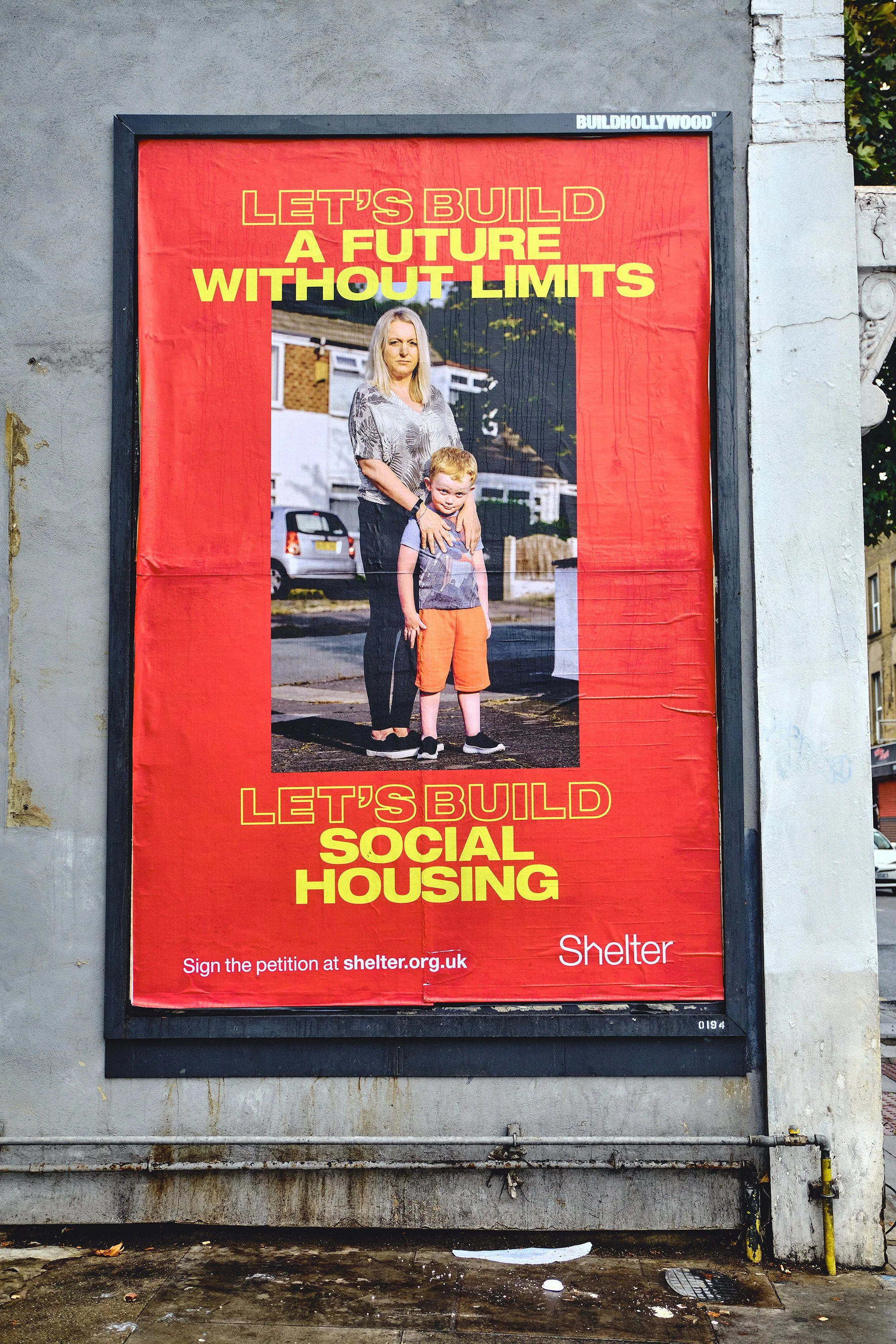 Red poster with a photo of a woman and a young boy standing on a street, promoting social housing with the text 'Let's build a future without limits' and 'Let's build social housing.'