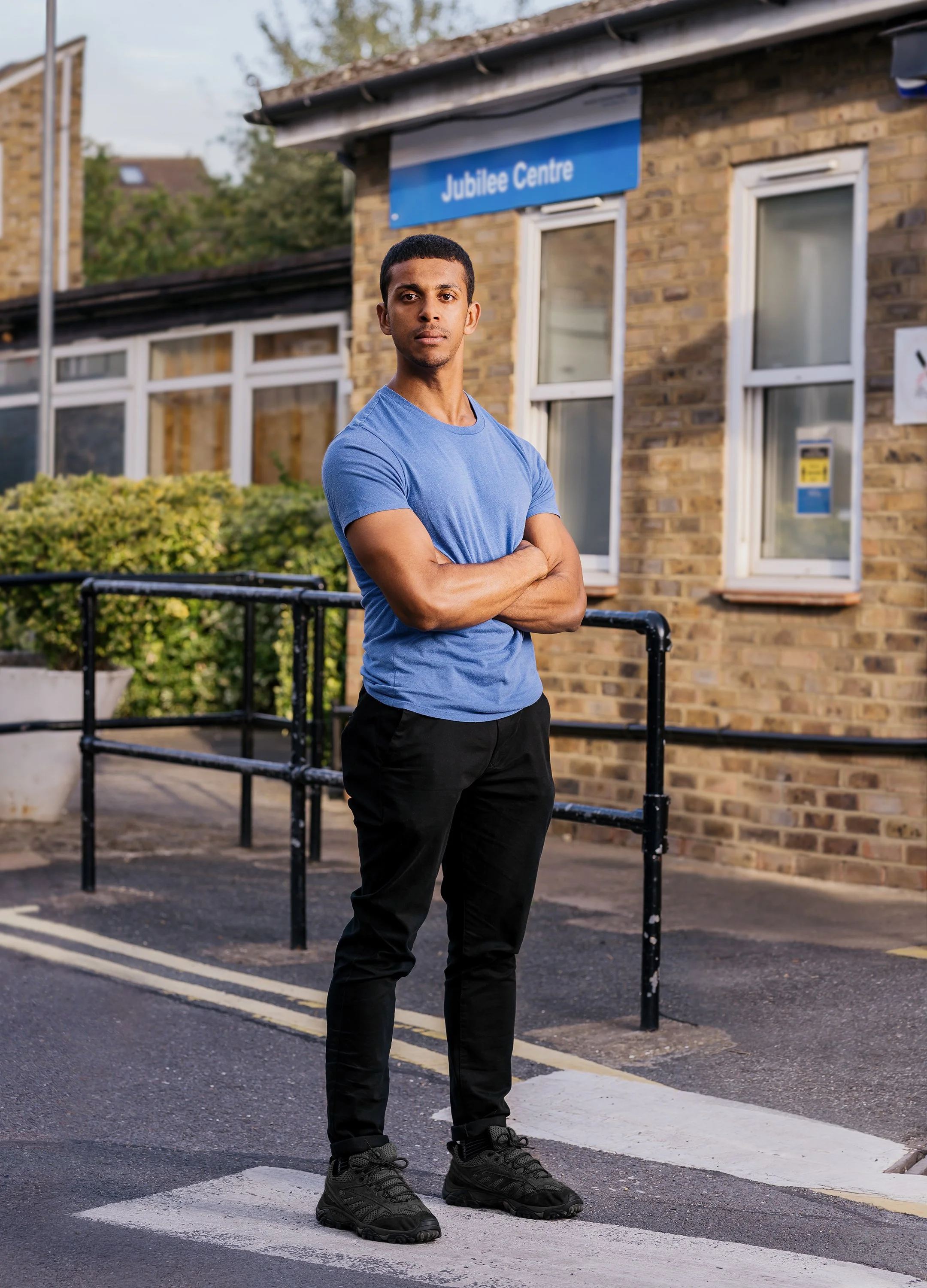 A young man in a blue t-shirt and black pants standing with arms crossed outside a brick Jubilee Centre building, with bushes and railings visible in the background, on a street with a crosswalk.
