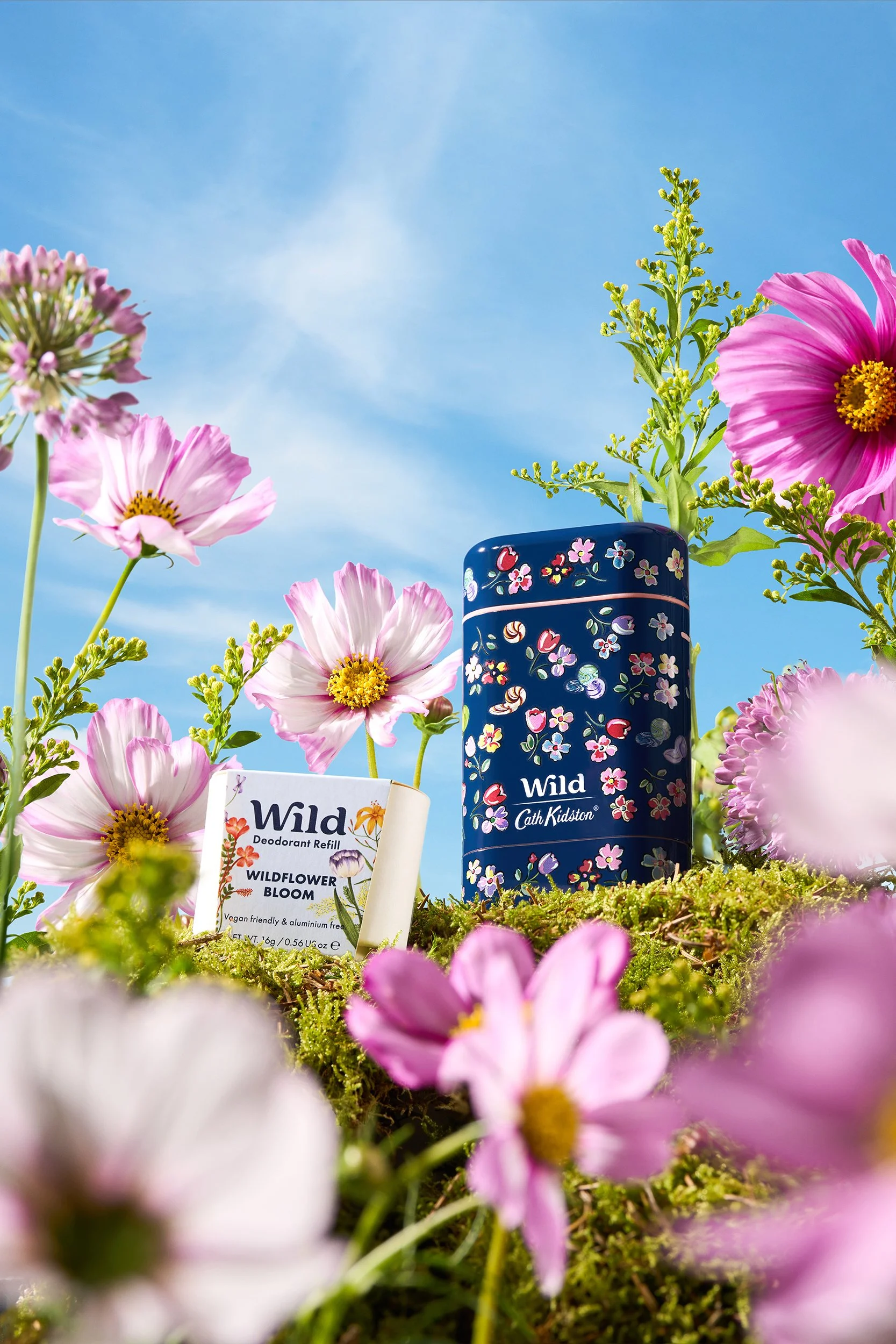 A floral display with pink and purple wildflowers surrounding a Wild deodorant refill and a blue floral-patterned tin container, set against a bright blue sky.