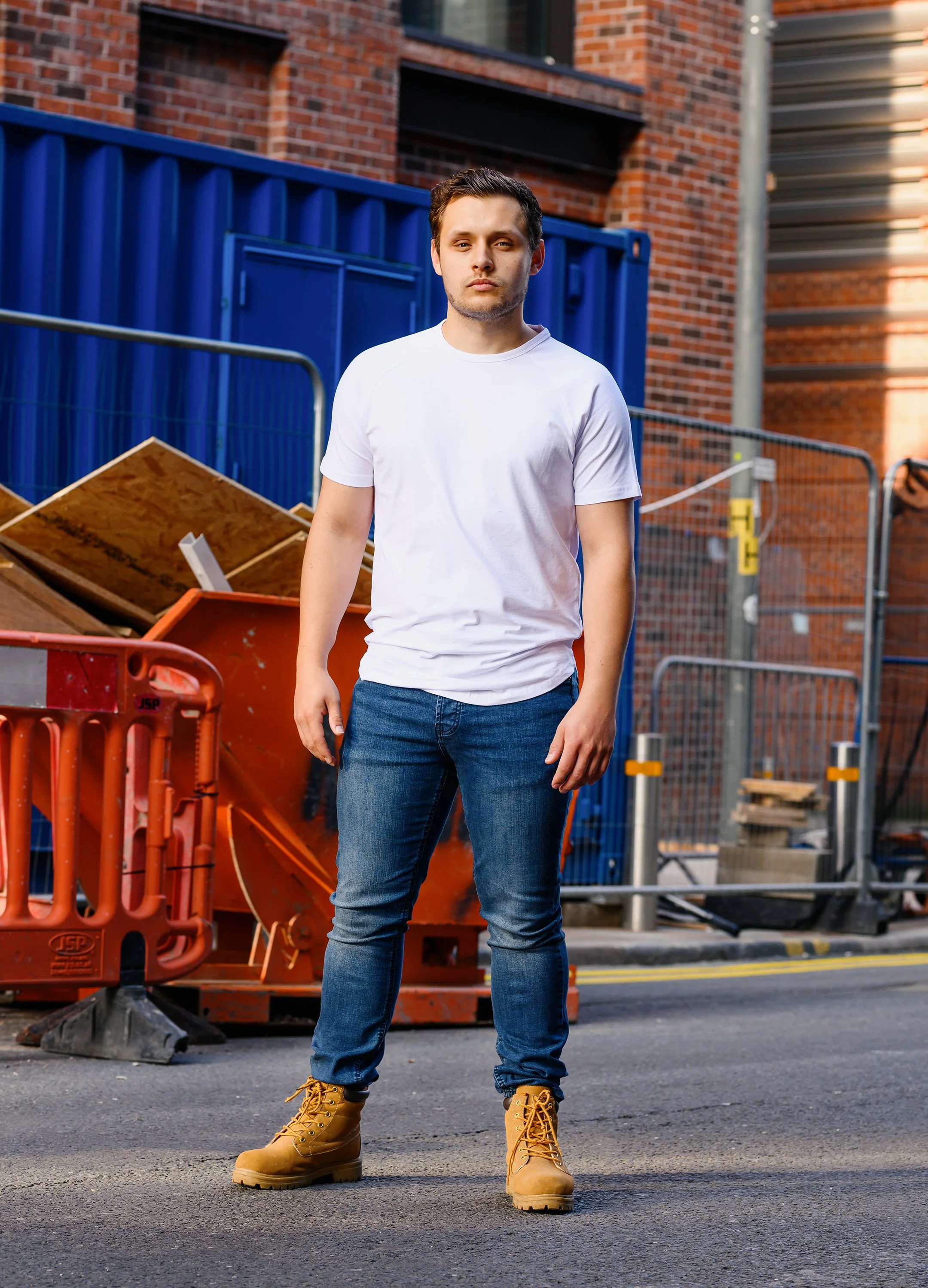 A young man standing on a city street in front of construction barriers and equipment, wearing a white t-shirt, blue jeans, and tan work boots.