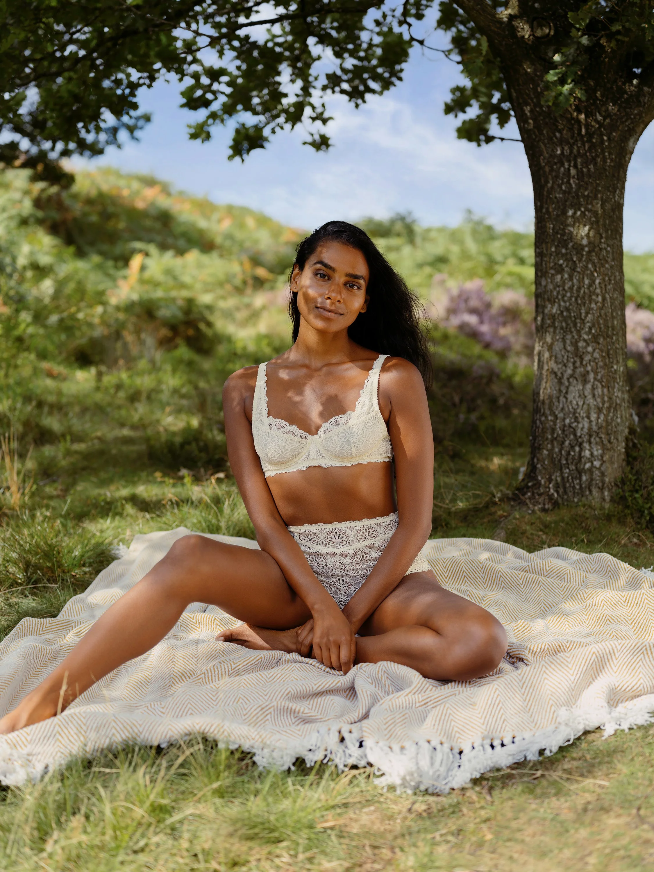 A woman in cream-colored lace lingerie sits on a patterned blanket under a tree outdoors, with green trees and a blue sky in the background.