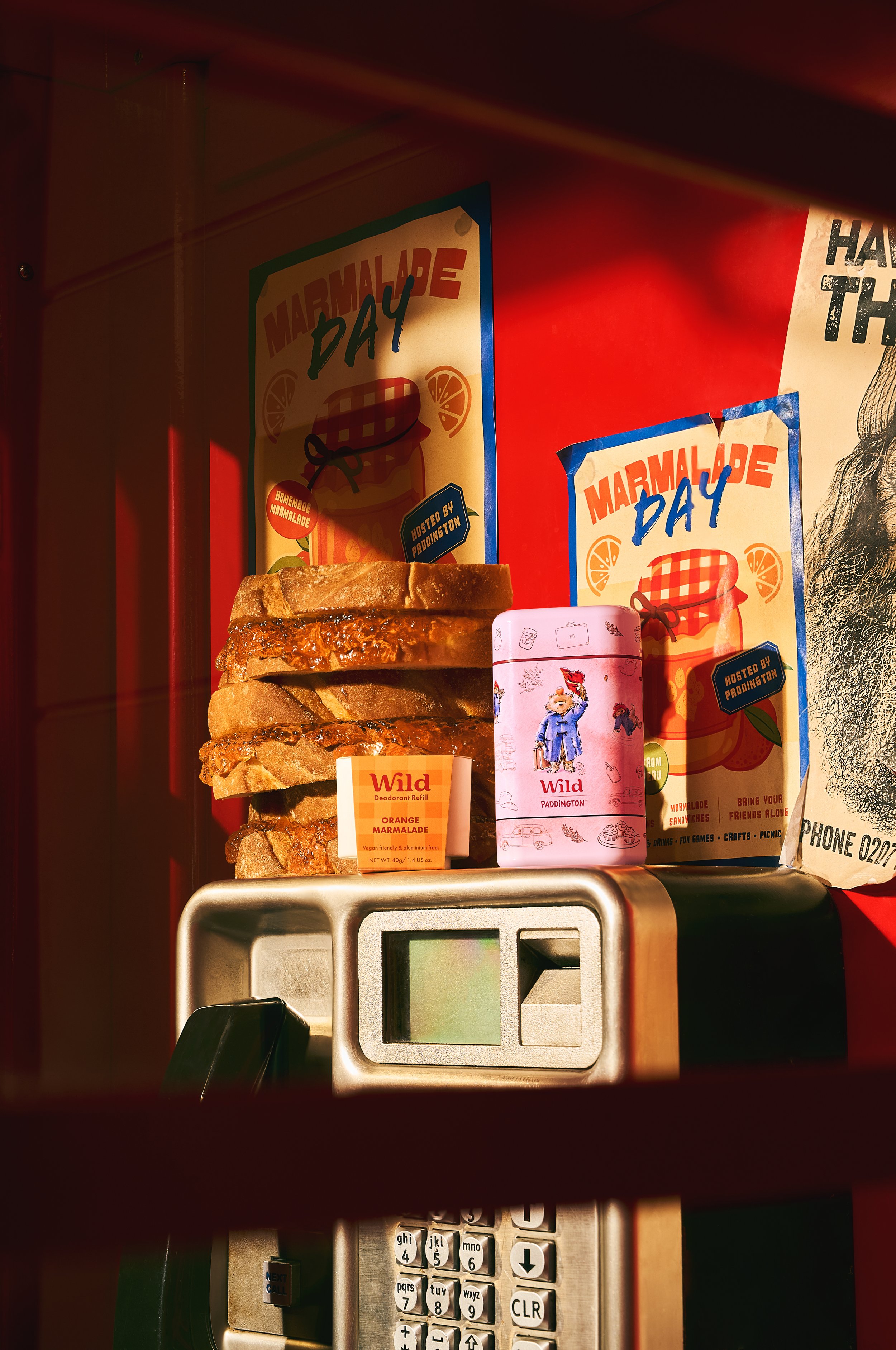 A vintage payphone with a stack of grilled bread slices, marmalade jars, and marmalade-themed posters on a red wall, illuminated by warm lighting.