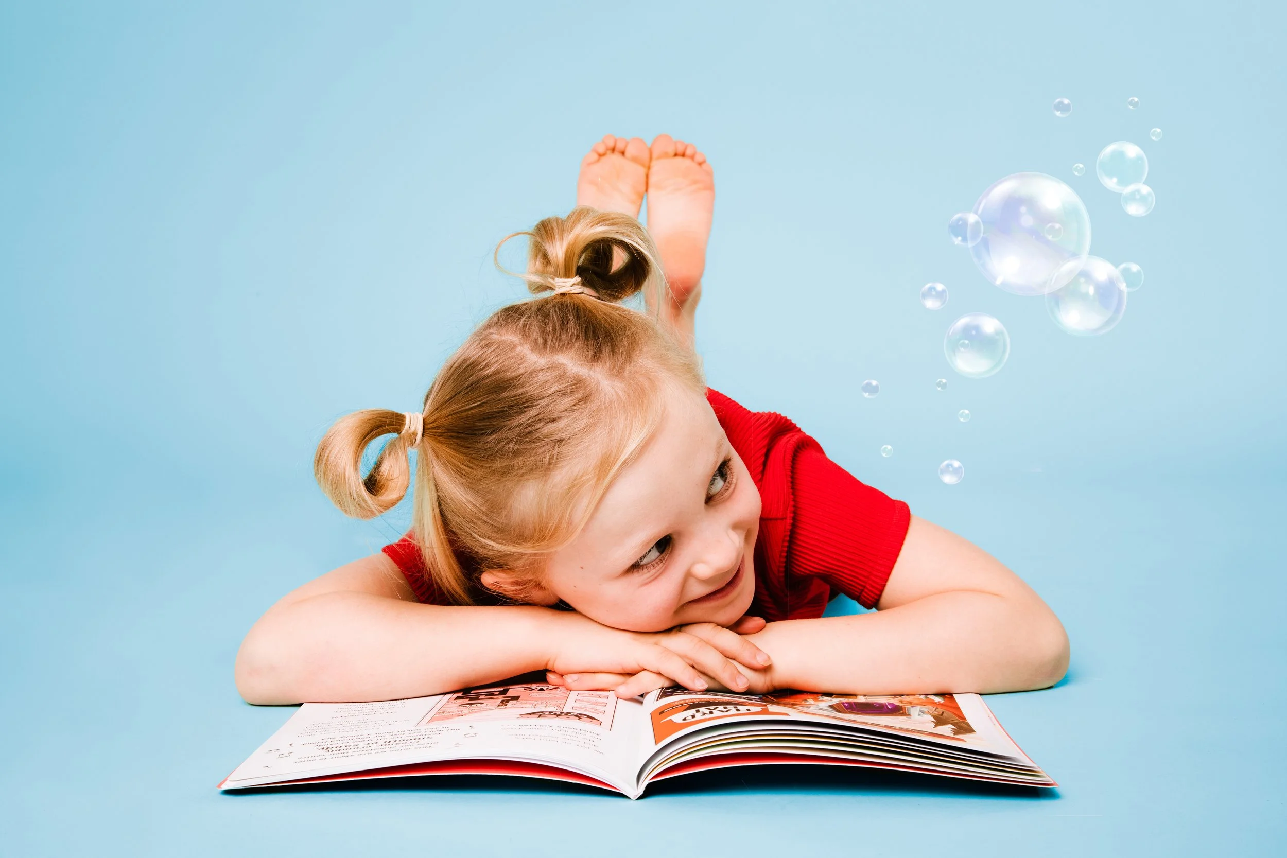 Two young girls with blonde hair, one with two ponytails and the other with a top knot, are lying on a flat surface looking at an open magazine against a light blue background. One girl is wearing a red shirt.