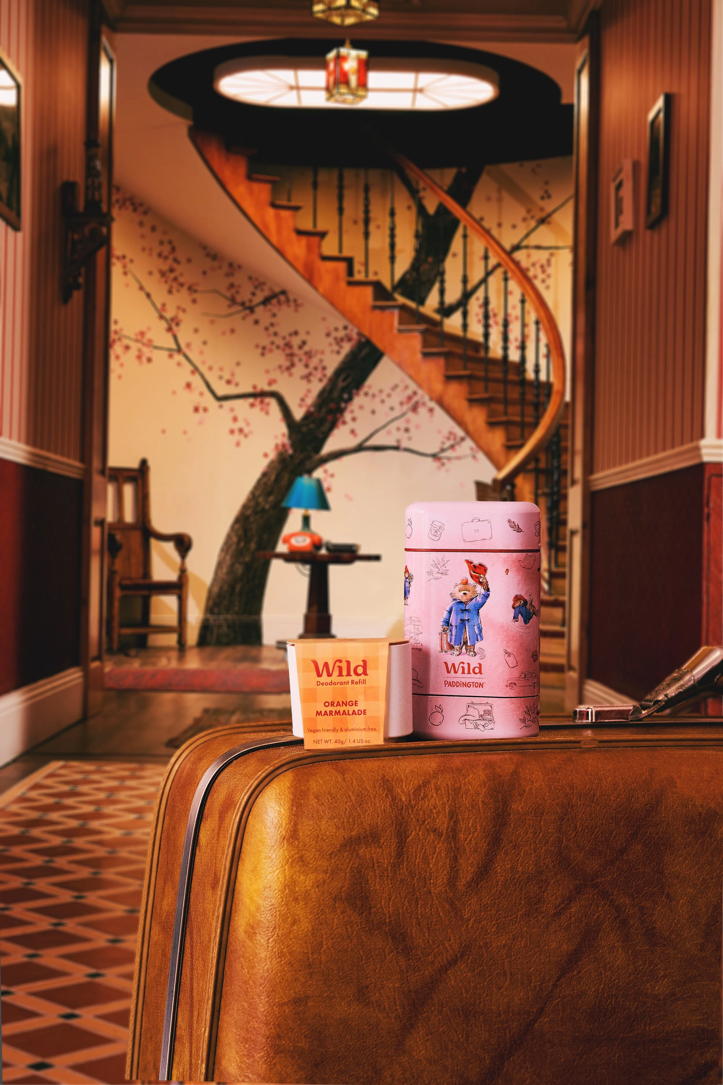A pink Tin of Paddington Bear themed tiles, a boxed refill of Wild deodorant with orange marmalade scent, and a small container of Wild deodorant refill, placed on a wooden table in a hallway with a staircase and a mural of a cherry blossom tree on the wall.