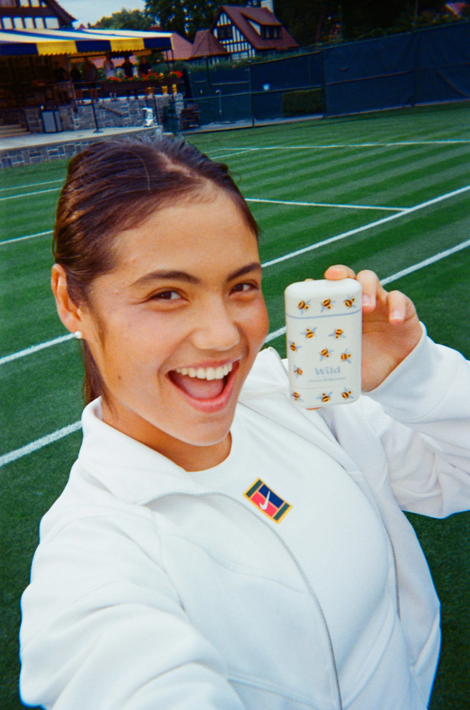 A young woman smiling and holding a small tin of Wild mints with bee illustrations, standing on a tennis court with houses and trees in the background.