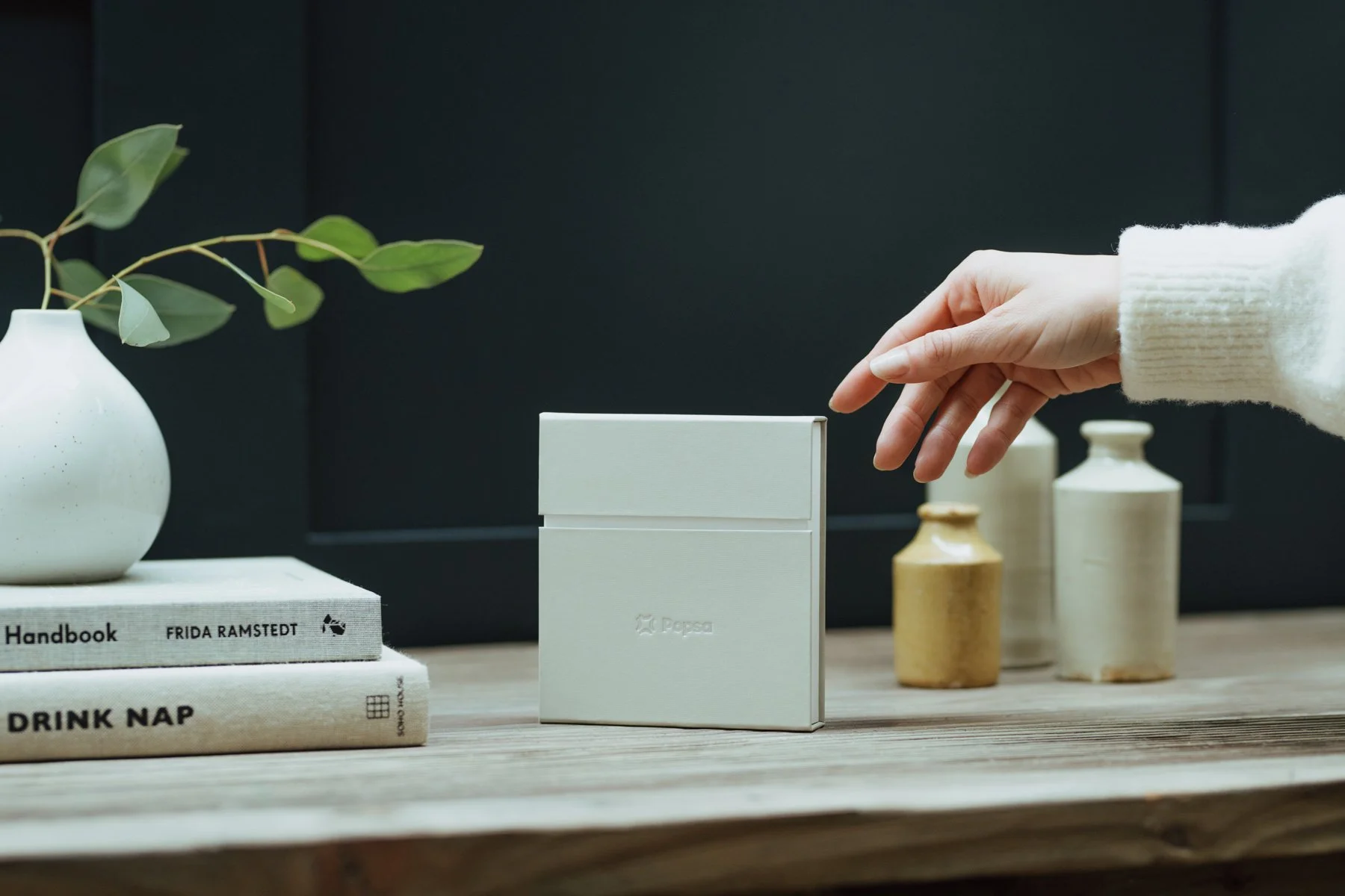A hand reaching toward a white box on a wooden table, with a stack of books, decorative vases, and a white vase with green leaves in the background.