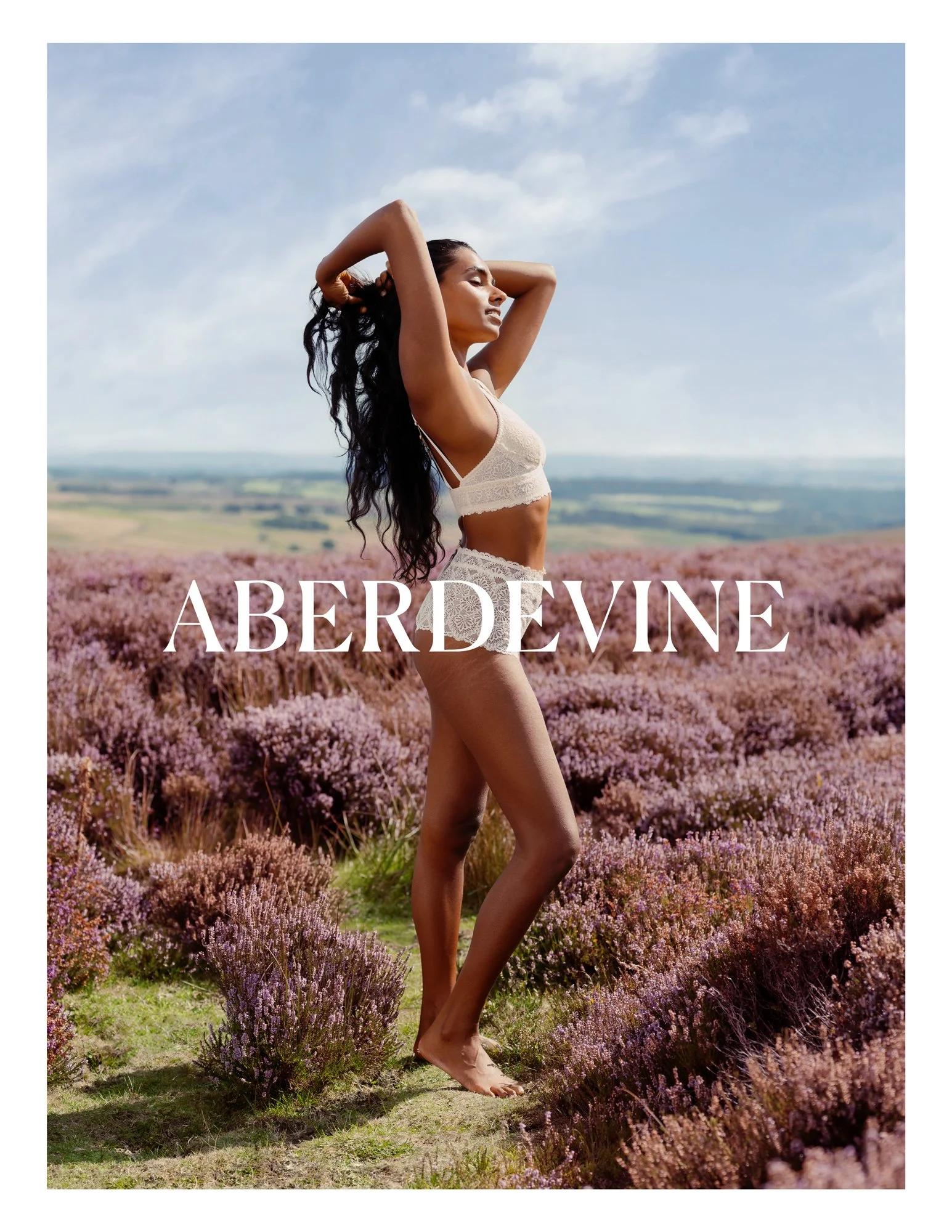 A woman standing in a field of purple flowers with her eyes closed, wearing a lace bralette and shorts, with her hands behind her head, against a backdrop of blue sky and distant rolling hills.