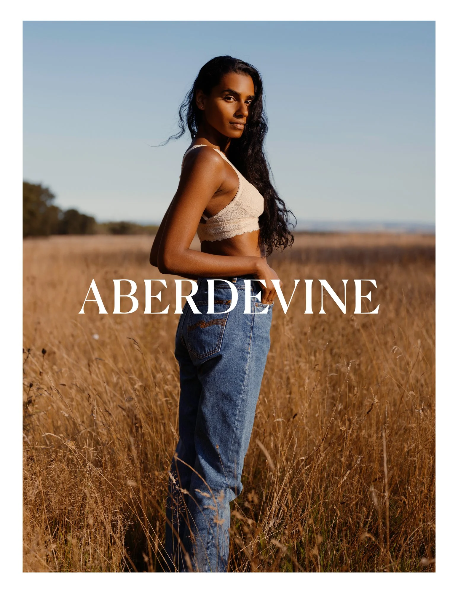 A woman with dark wavy hair and a tan complexion standing in a field of tall dry grass under a clear blue sky, wearing a beige lace crop top and high-waisted blue jeans.