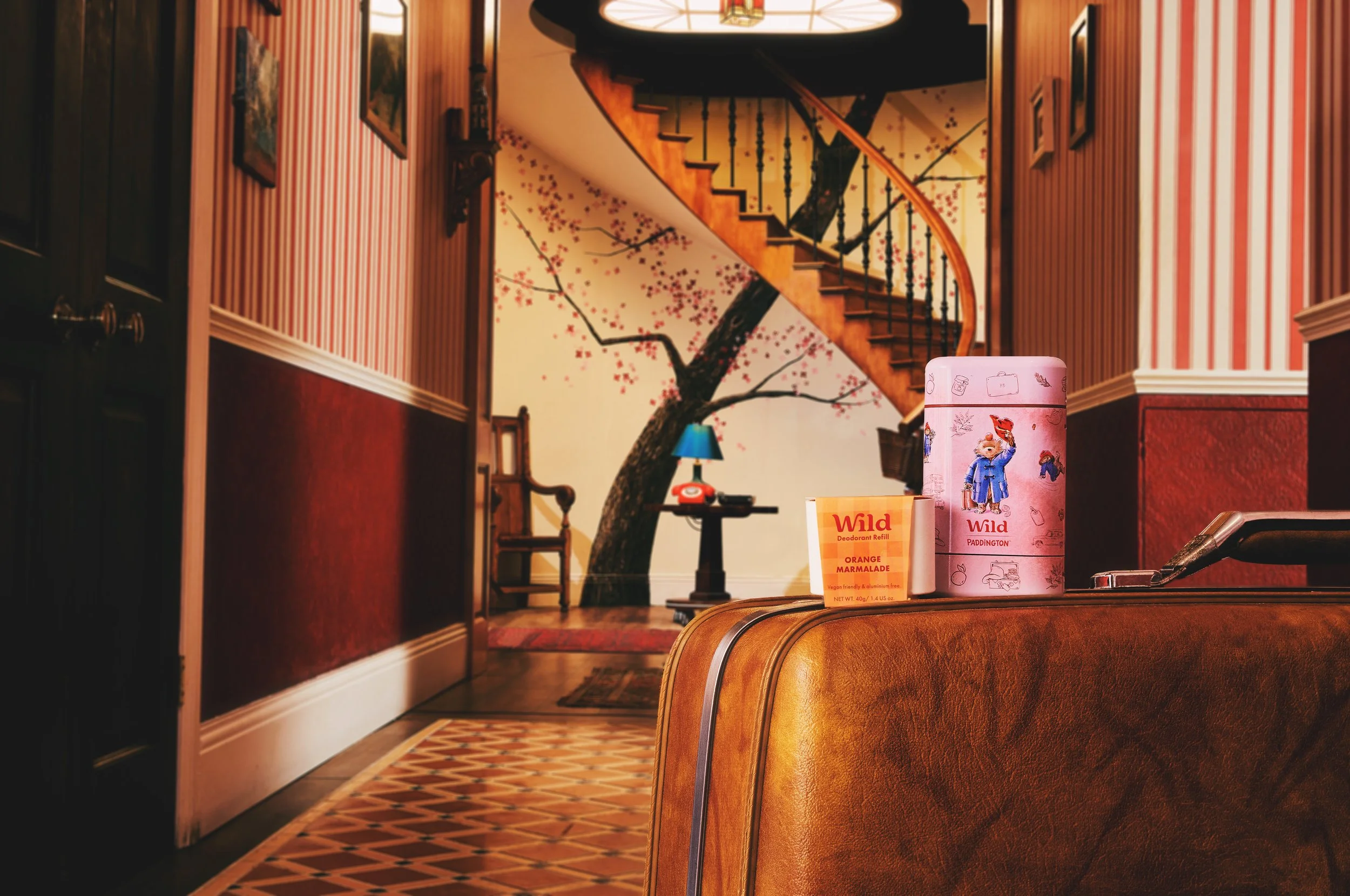 Interior view of a house with vintage furniture, including a suitcase and decorative items on top. The background features a staircase with a curved wooden banister and a painted mural of a cherry blossom tree on the wall. There is a small table with a blue lamp and a chair near the wall.